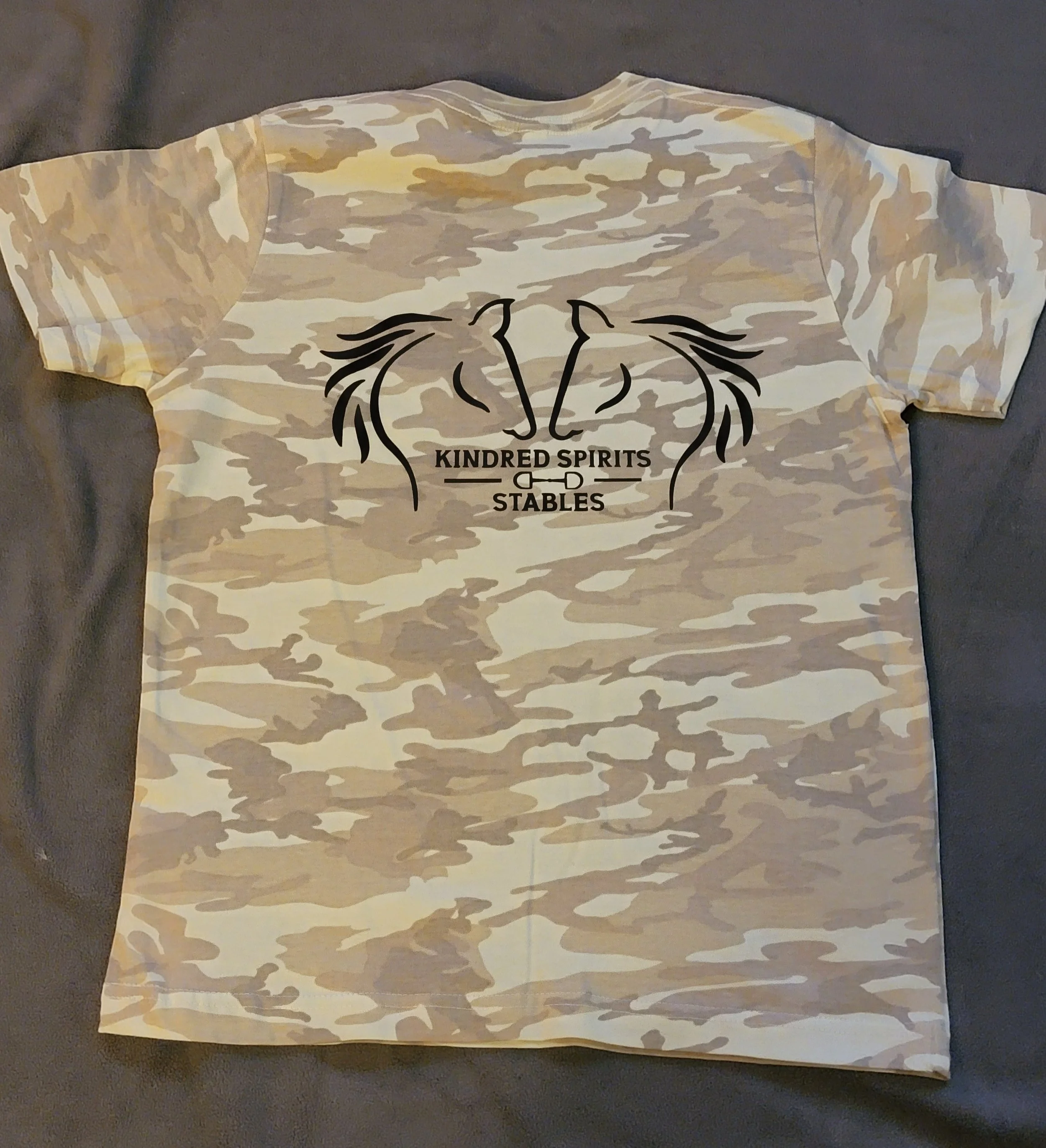 Camouflage-patterned T-shirt with a black graphic of two horses facing each other, a sword through the middle, and the text "KINDRED SPIRITS STABLES" beneath.