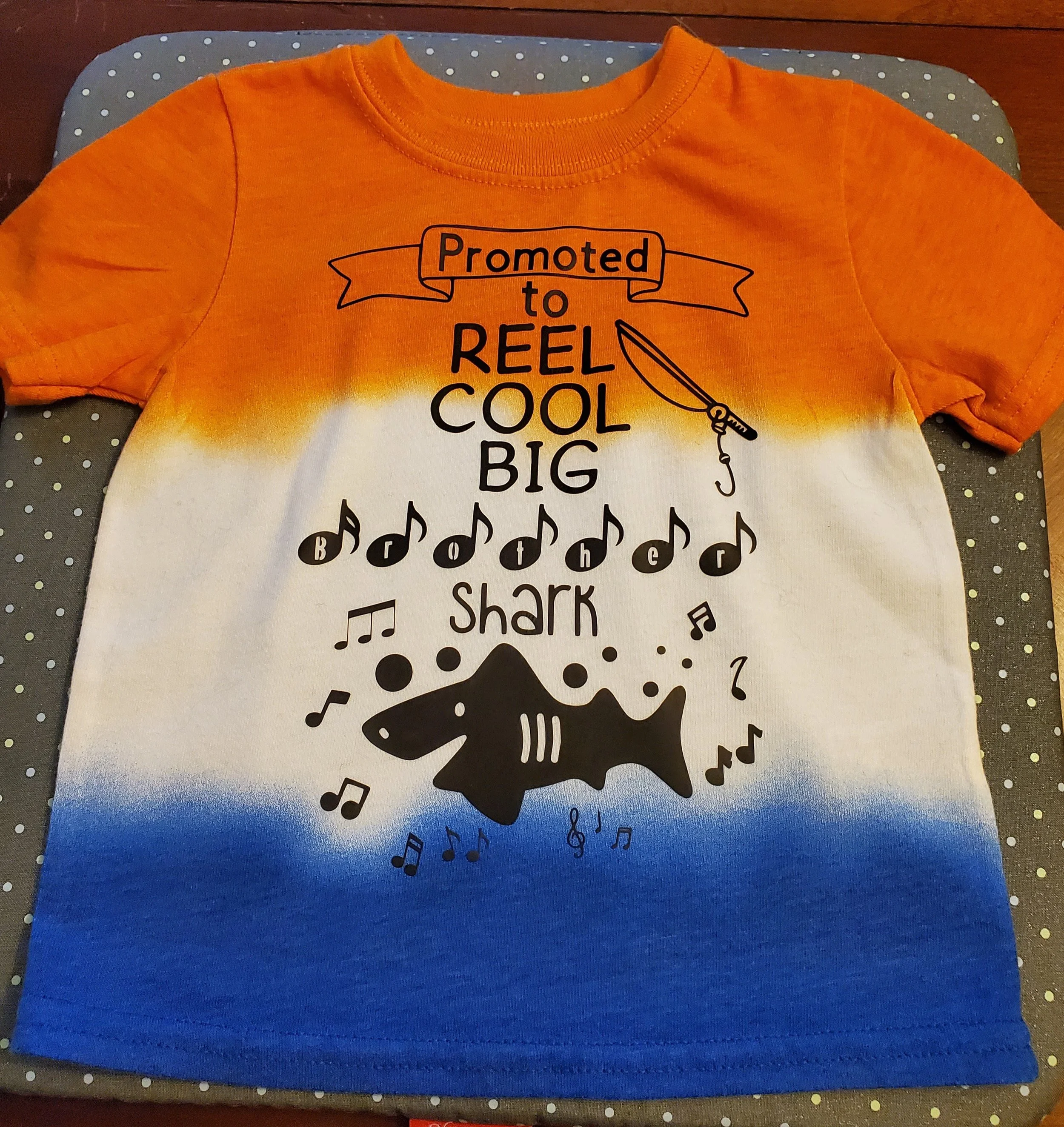 A children's T-shirt with a gradient orange, white, and blue background. It features a fishing rod, musical notes, and a shark graphic. The text reads 'Promoted to Reel Cool Big Shark'.