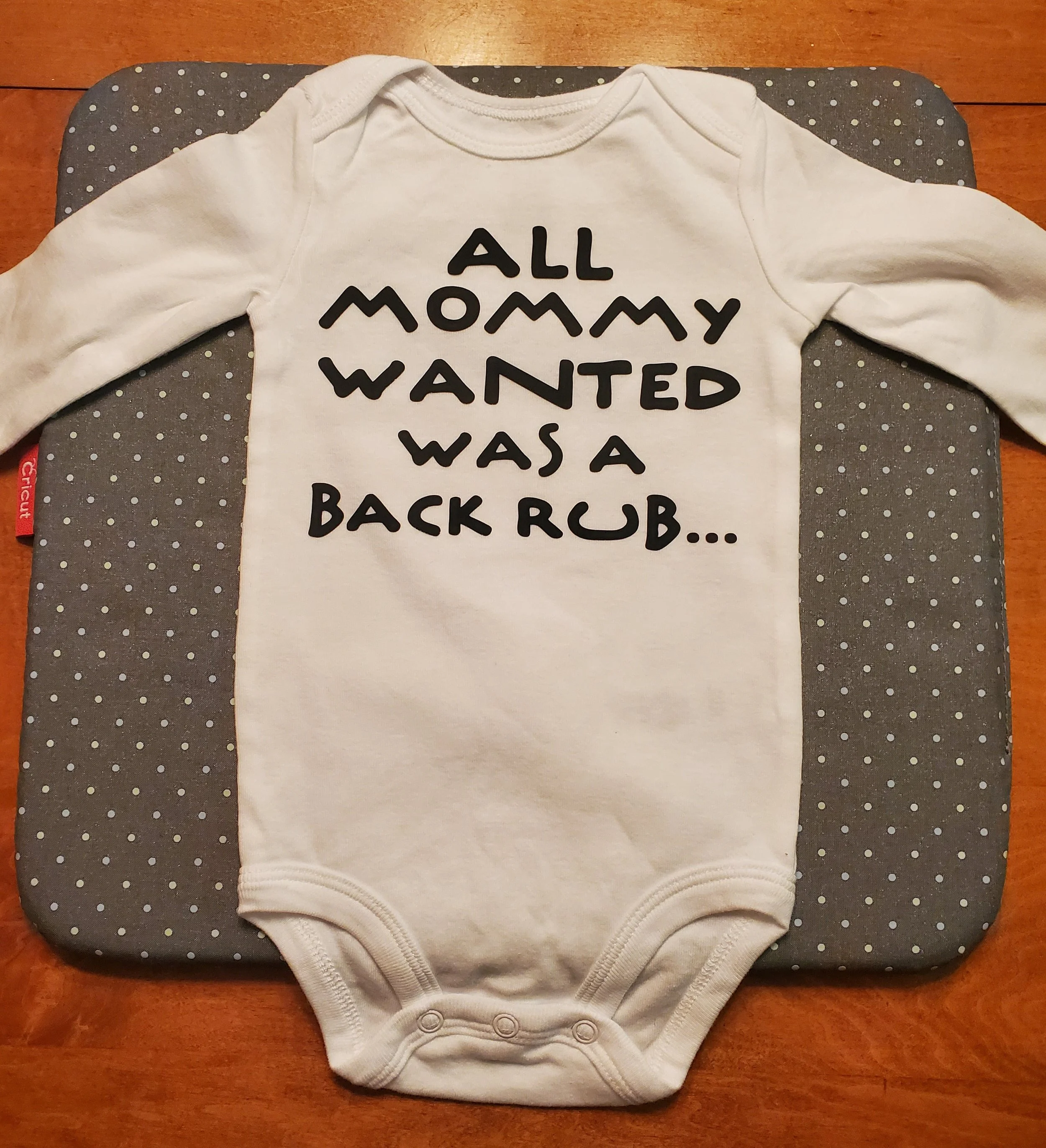 Baby onesie with text reading "All mommy wanted was a back rub" laid flat on a polka dot changing pad on a wooden surface.