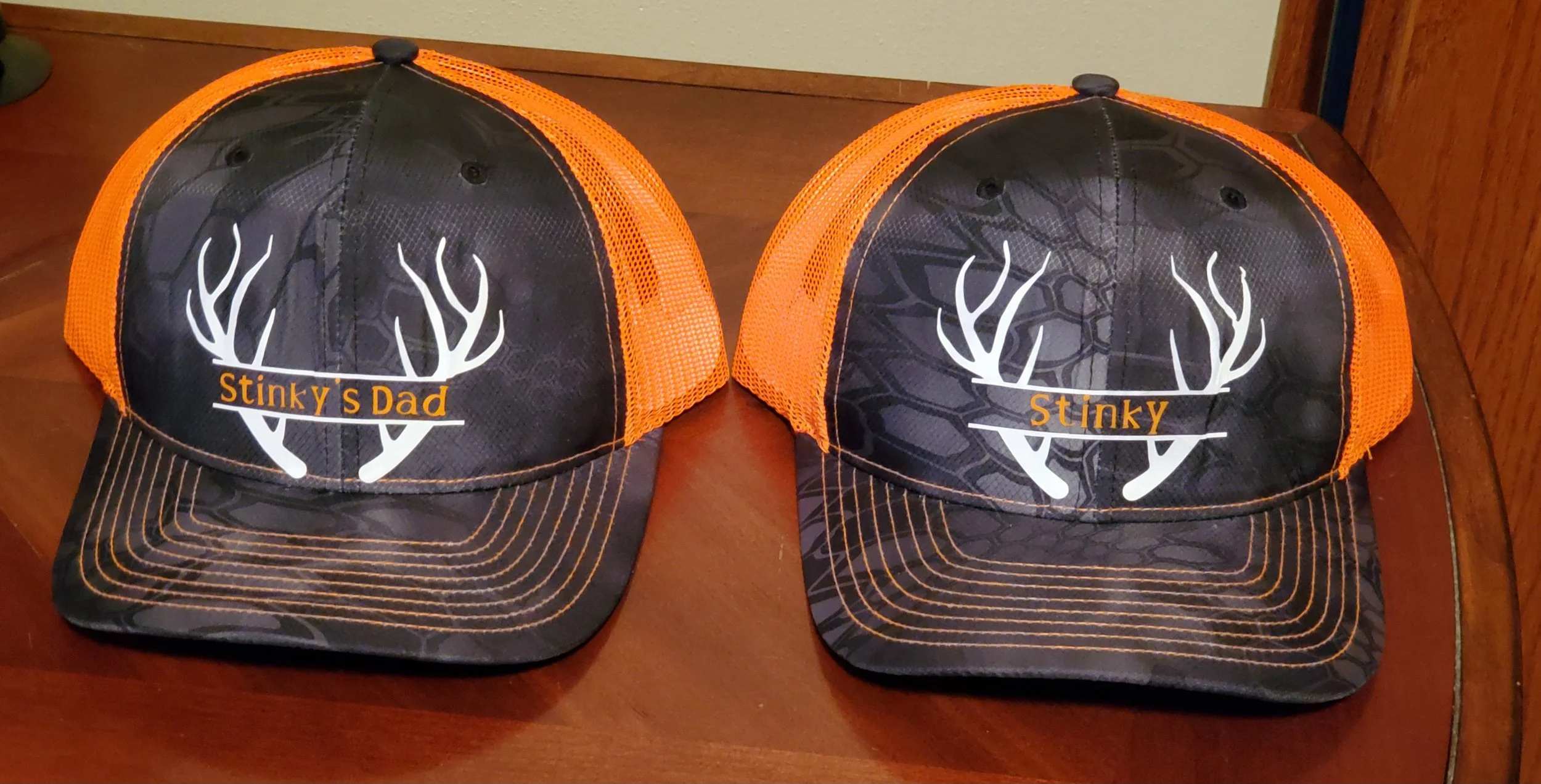 Two black and orange trucker hats with a white deer antlers graphic on the front and the words 'Stinky s Dad' and 'Stinky' written in orange.