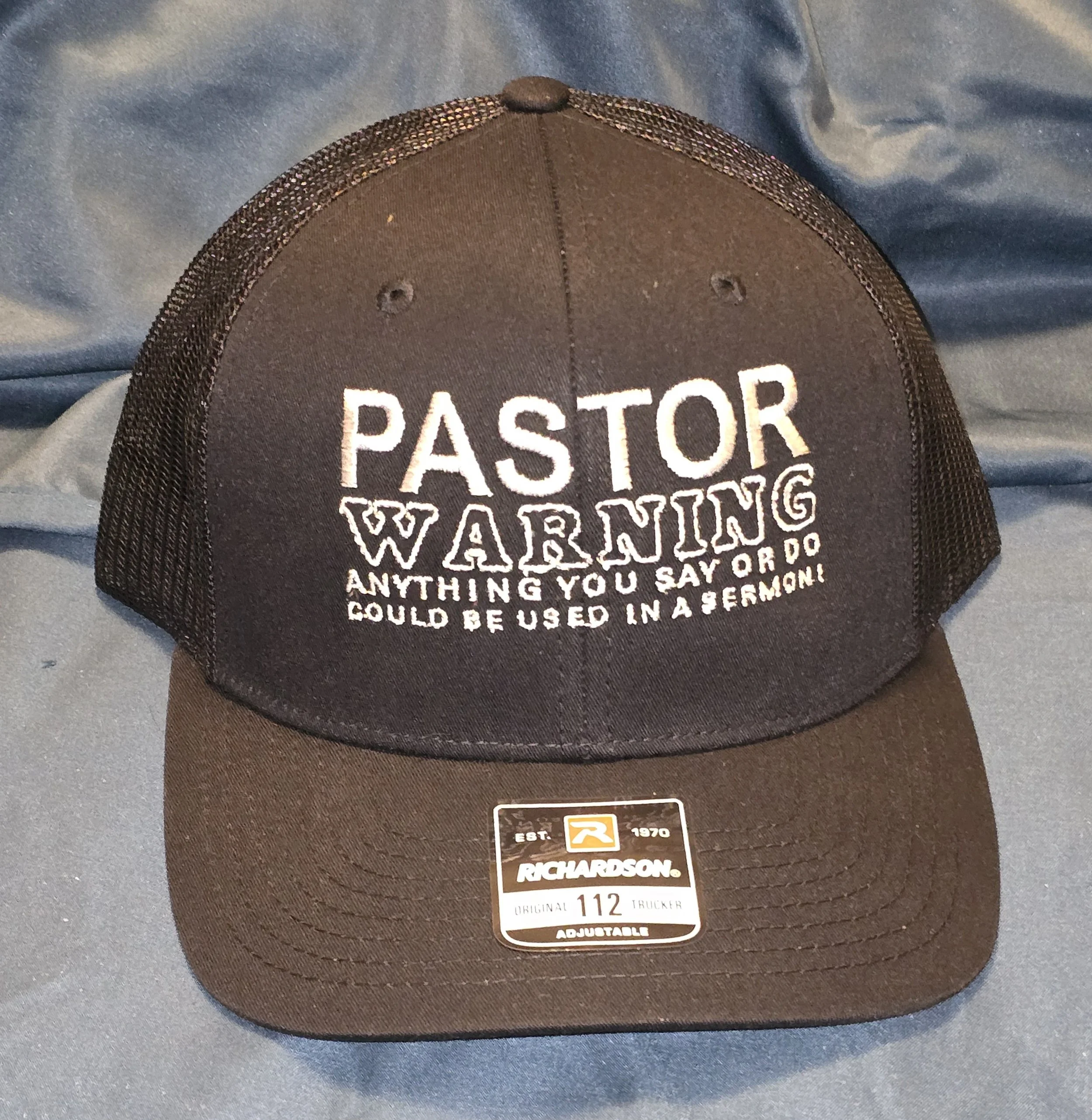 Black trucker cap with white embroidered text reading 'Pastor Waring Anything You Say Or Do Could Be Used in a Sermon!'. The cap has a mesh back and a sticker on the brim that says 'Richardson 112 Original Trucker Adjustable'.