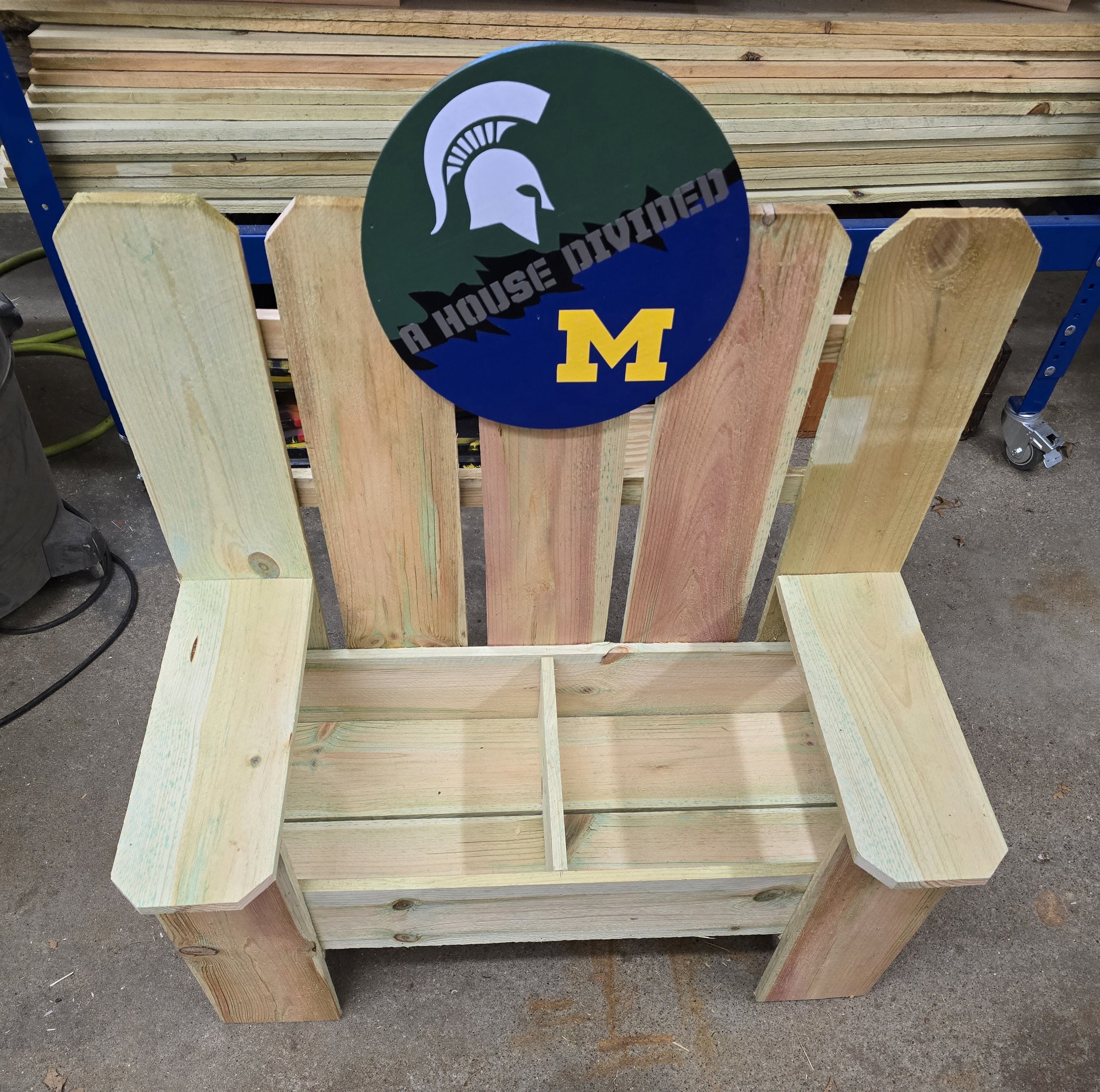 A wooden bench with a Michigan State Spartans logo and a University of Michigan logo attached to the backrest, with a sign saying 'A House Divided' in the middle.