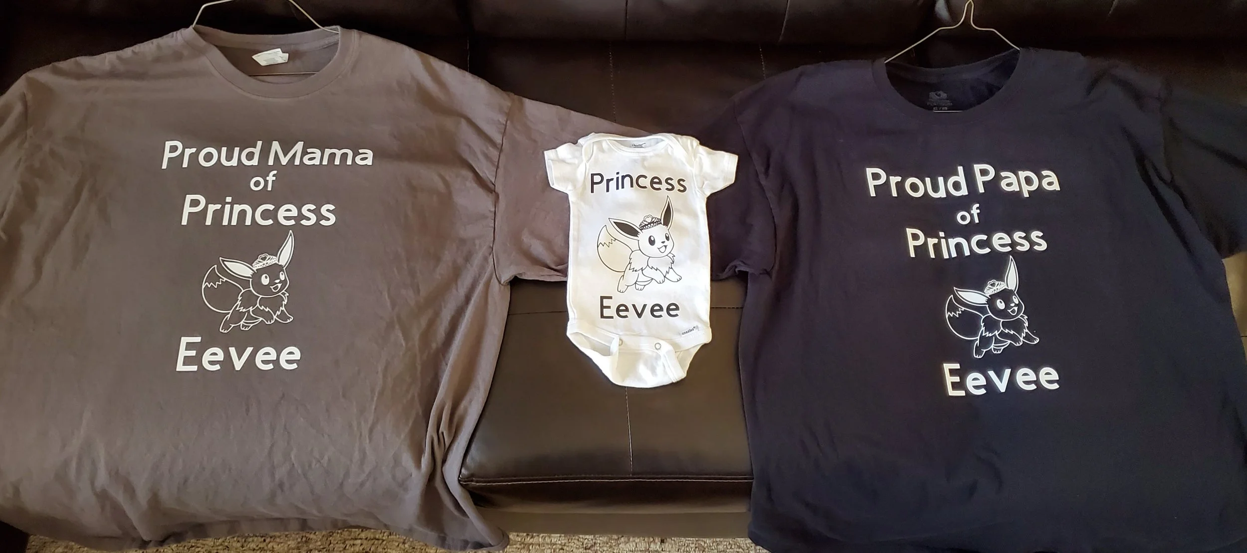 Three clothing items with matching designs: a gray T-shirt reading 'Proud Mama of Princess Eevee,' a white onesie reading 'Princess Eevee' with a cute cartoon Eevee wearing a crown, and a black T-shirt reading 'Proud Papa of Princess Eevee,' all feat