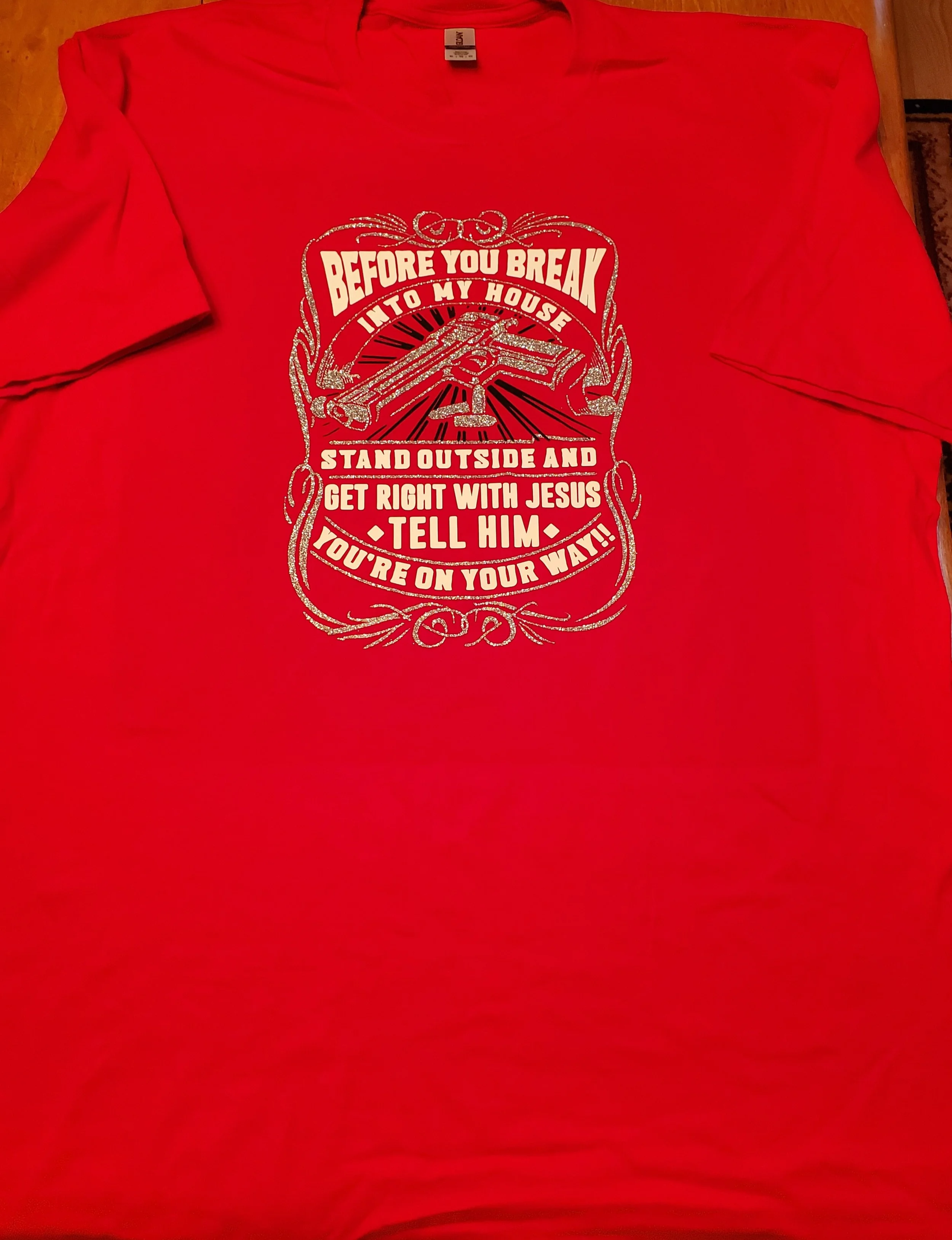 Red T-shirt with a graphic design that includes a gun, rays, and decorative elements, along with the words: "BEFORE YOU BREAK INTO MY HOUSE, STAND OUTSIDE AND GET RIGHT WITH JESUS, TELL HIM, YOU'RE ON YOUR WAY!!"