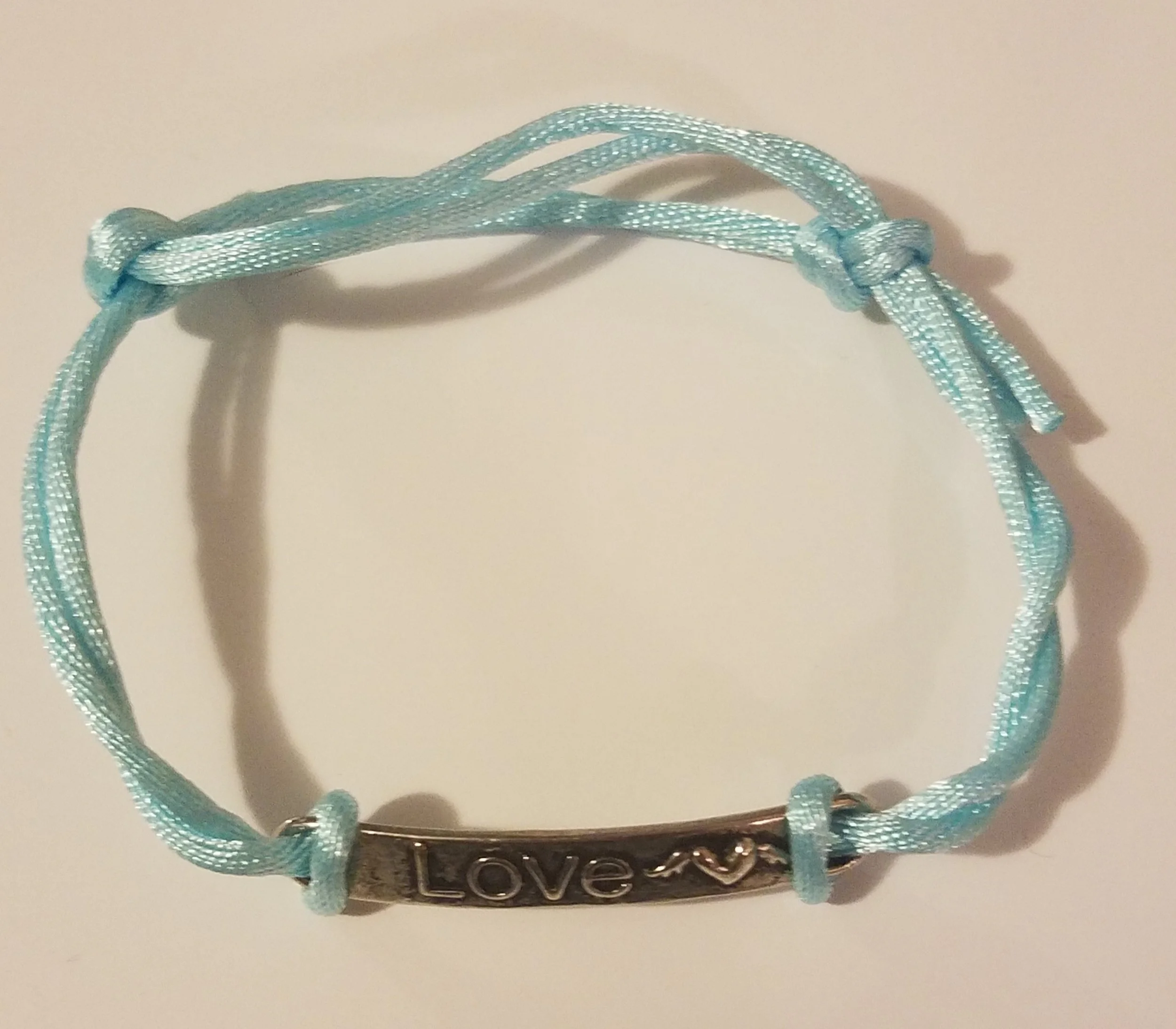 Light blue braided cord bracelet with a metal plate engraved with the word 'Love' and a heart symbol in the middle.
