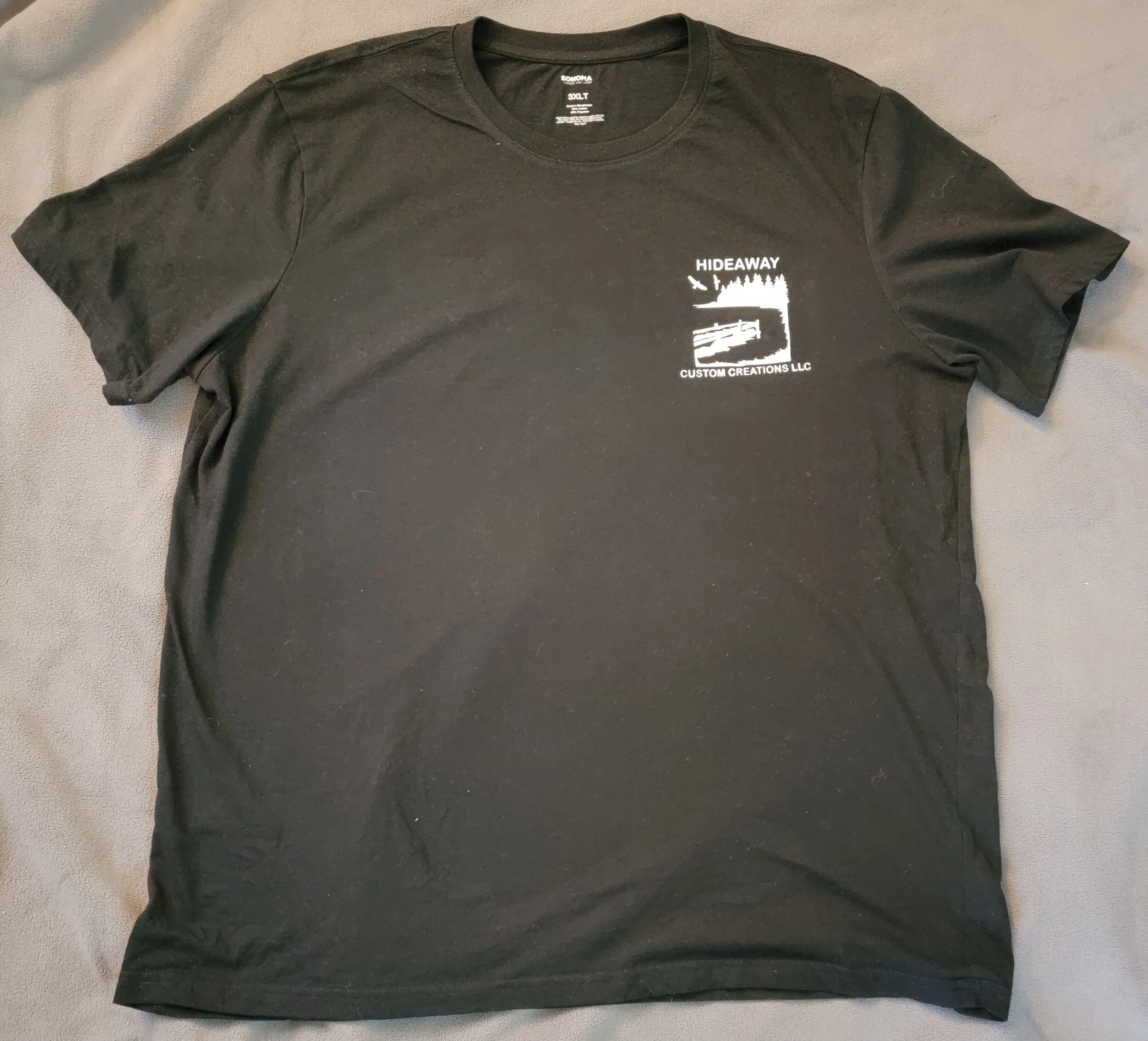 Black T-shirt with white graphic and text on the upper left side reading "HIDEAWAY CUSTOM CREATIONS LLC," featuring an outline of a landscape scene with trees, water, and a flying bird.