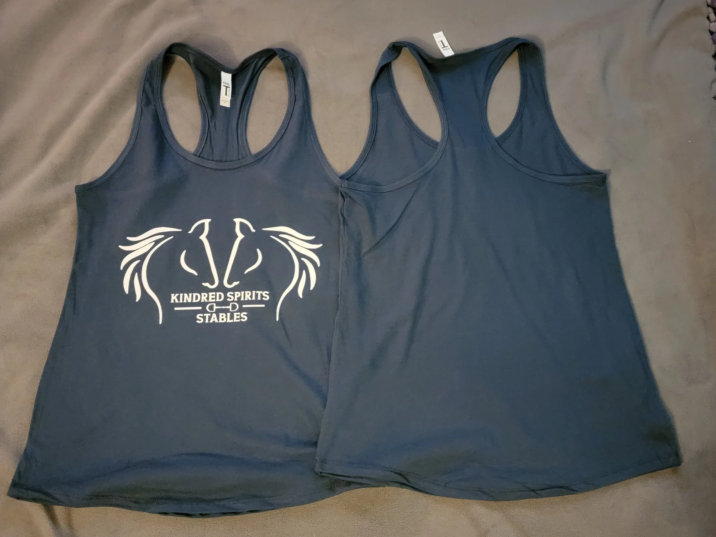 Two navy blue tank tops laid flat on a dark surface, one with a white logo that reads 'Kindred Spirits Stables' along with a horse head design.
