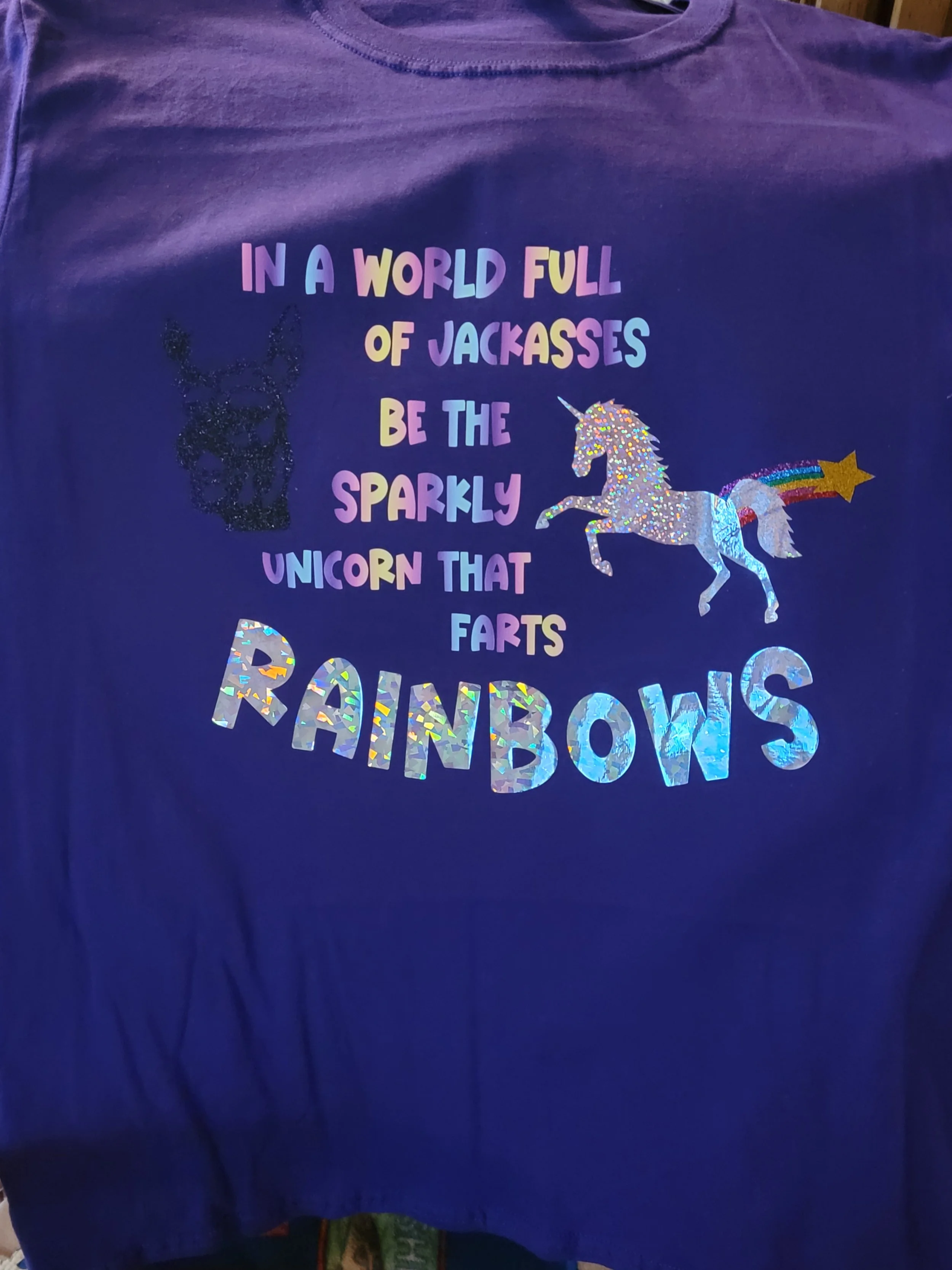 Blue T-shirt with colorful glittery text and images of a unicorn and a dog. The text reads "In a world full of jackasses be the sparkly unicorn that farts rainbows." The unicorn is decorated with glitter and has a rainbow trail.