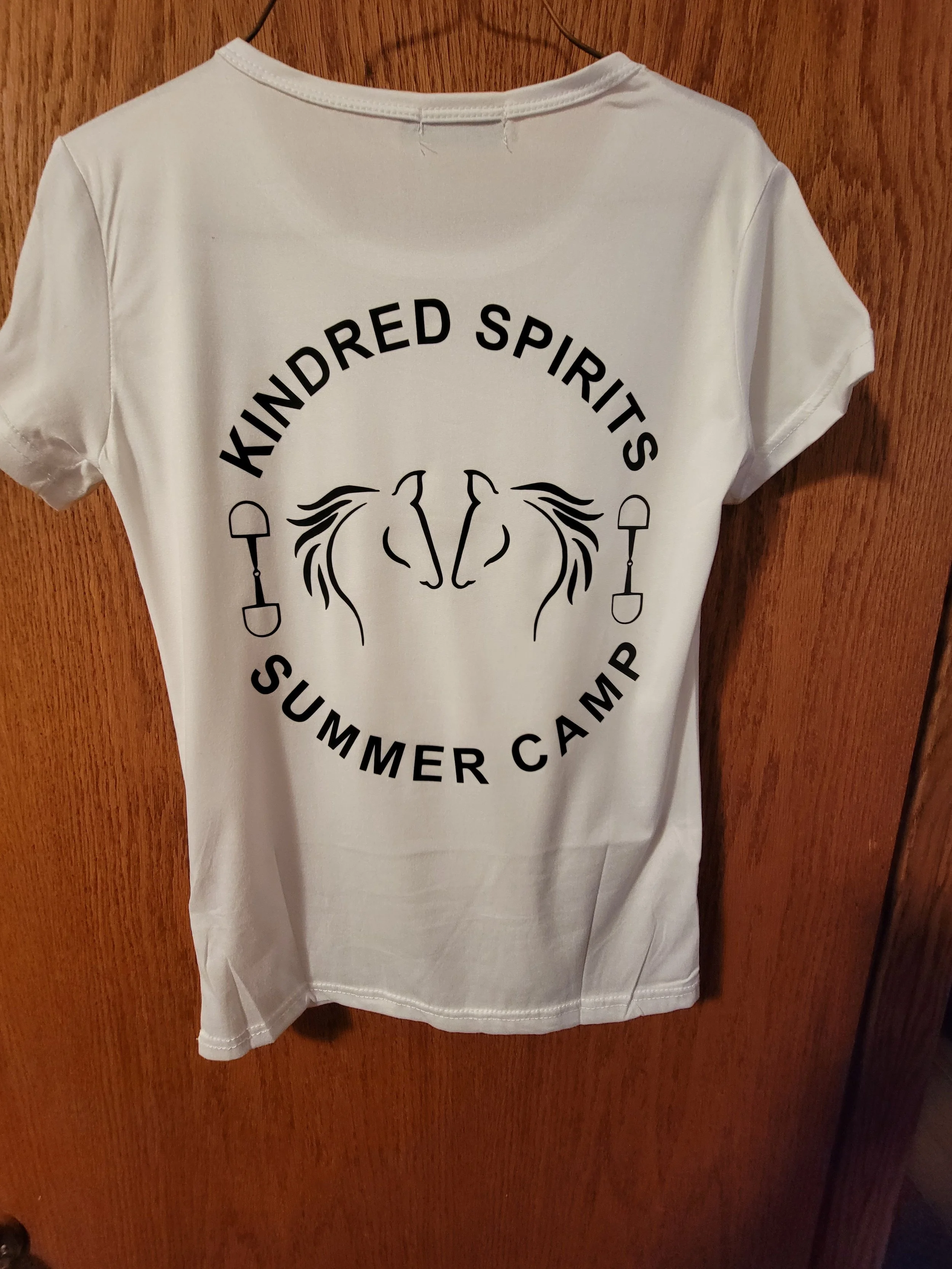 White t-shirt hanging on a wooden door with black text and a graphic design that reads 'Kindred Spirits Summer Camp.' The design features two line-drawn horses facing each other with their manes intertwined, flanked by two small shovels.