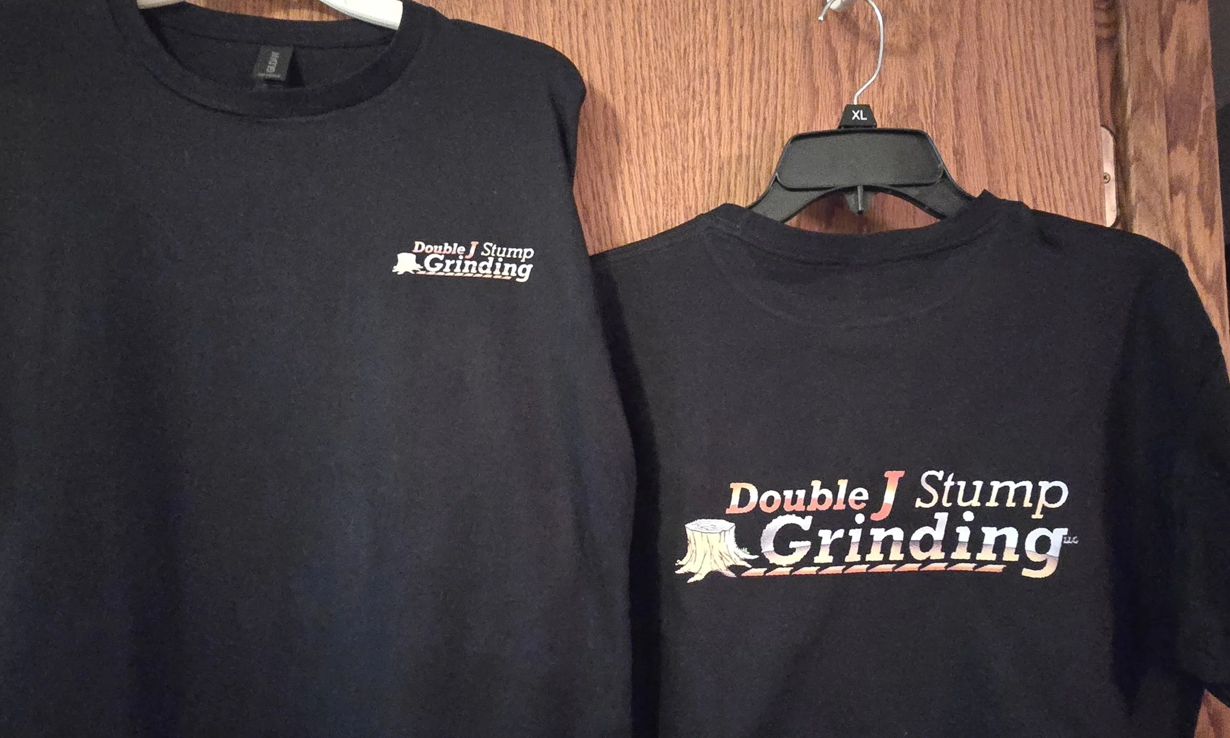 Two black T-shirts hanging on a wooden wall, one on the left with small logo, one on the right with large logo. The large logo has the text 'Double J Stump Grinding' with an illustration of a tree stump.