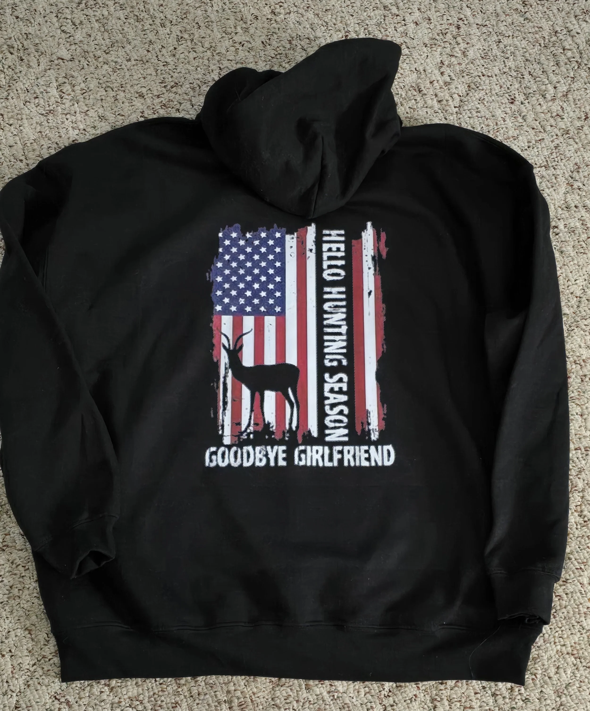 Black hoodie with a distressed American flag print, a silhouette of a goat, and the text 'Goodbye Girlfriend' and 'Hello Hunting Season' printed on the back, lying on a beige carpet.