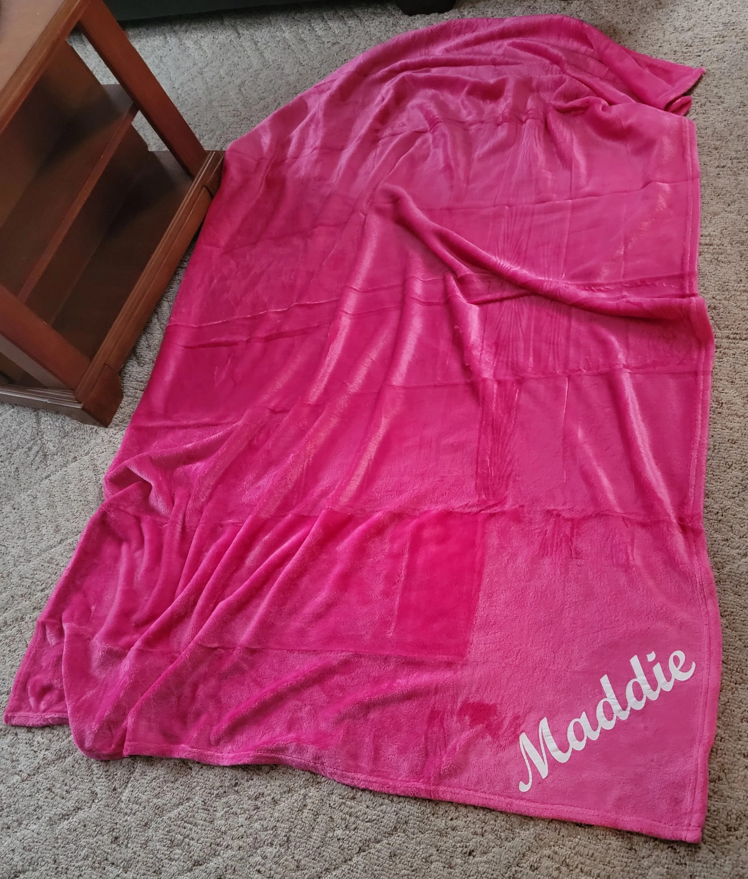 Pink plush blanket with the name 'Madie' written in white script near the corner, spread out on a beige carpet next to a wooden bookshelf.