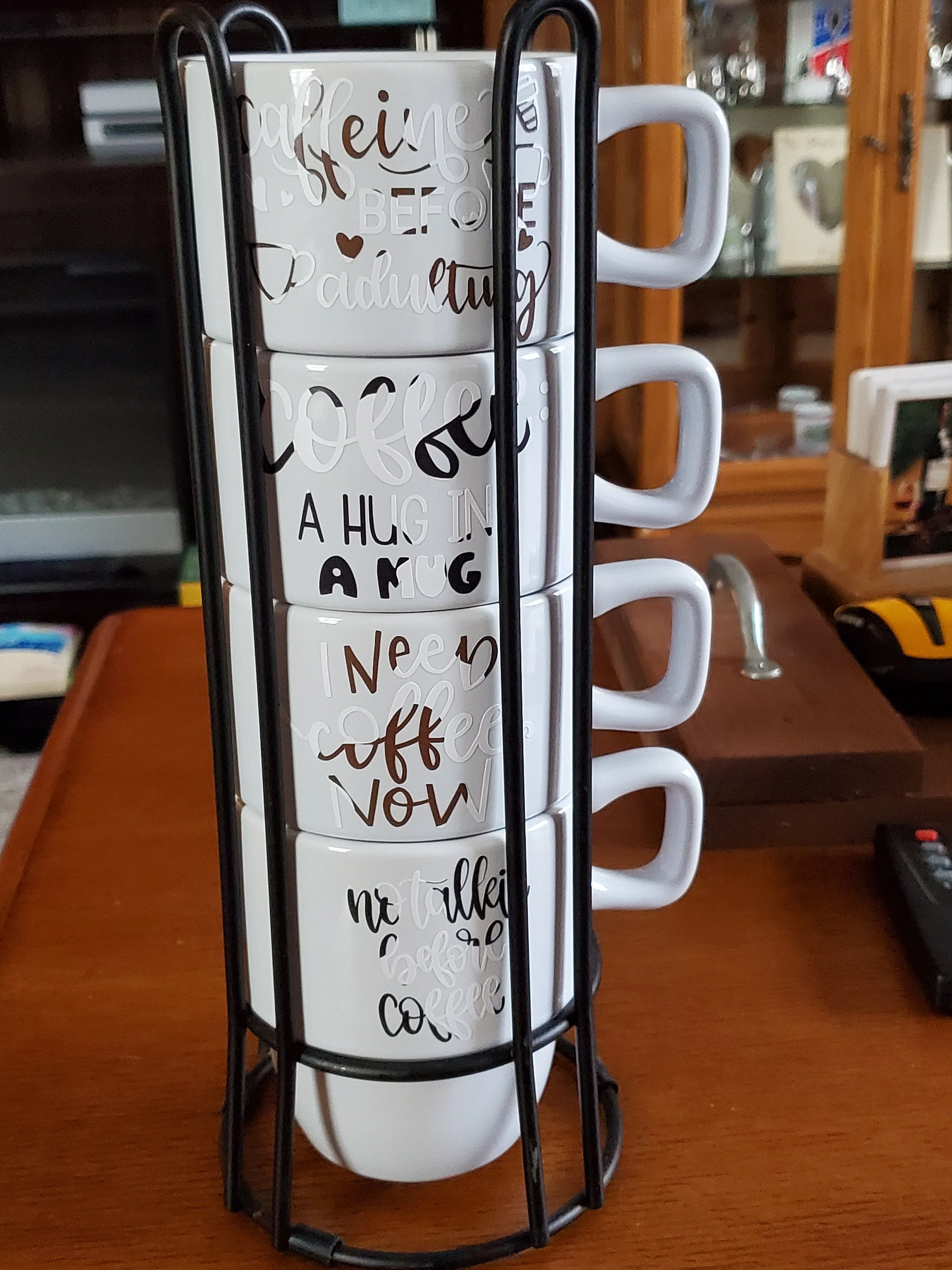 Four white coffee mugs with different black and brown text designs, stacked in a black wire mug holder on a wooden table.