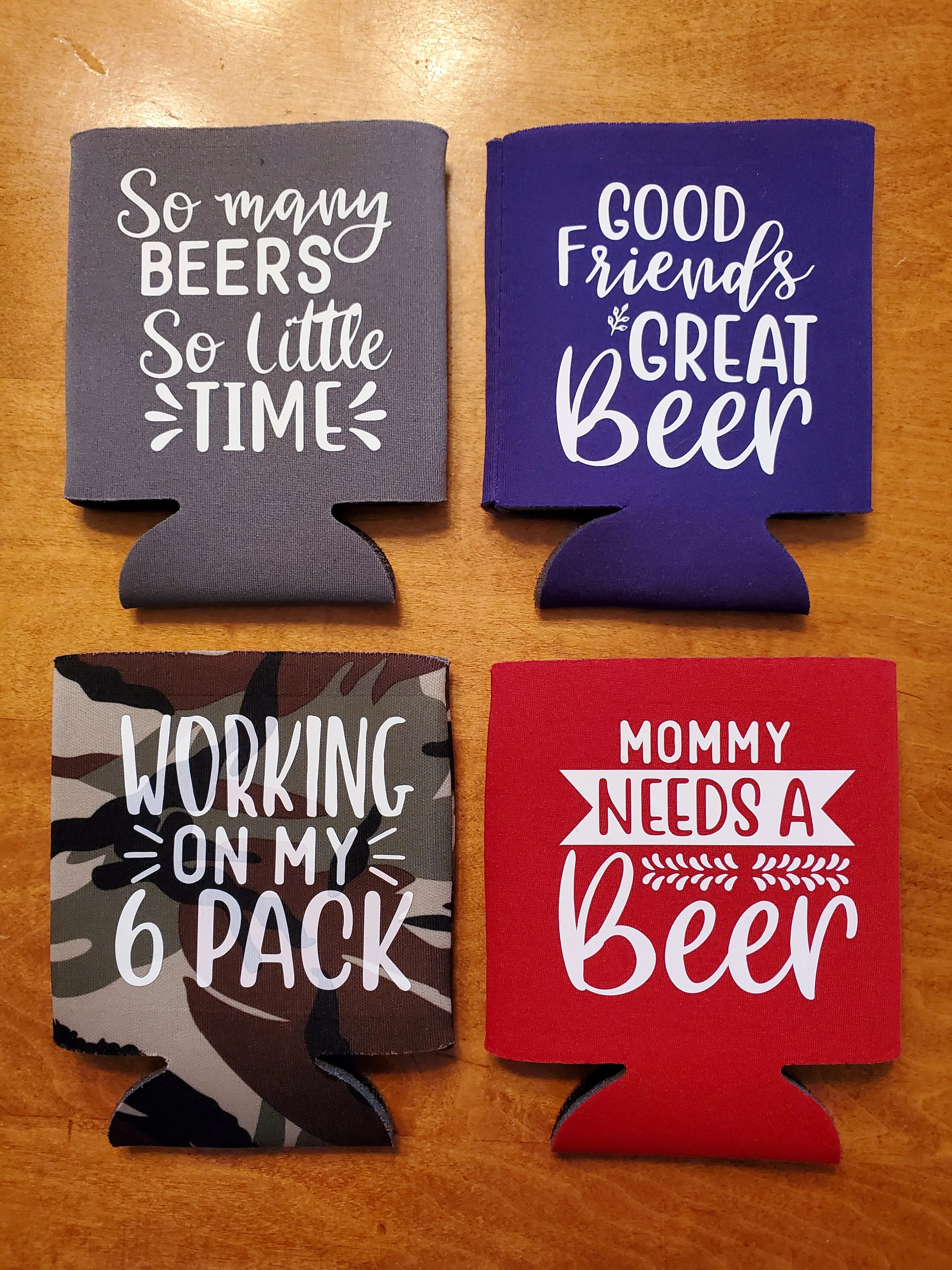 Four beverage coasters with humorous messages about beer, arranged in a 2x2 grid on a wooden surface. Top left is gray with white text, saying "So many BEERS So little TIME." Top right is blue with white text, saying "GOOD Friends & GREAT Beer." Bott