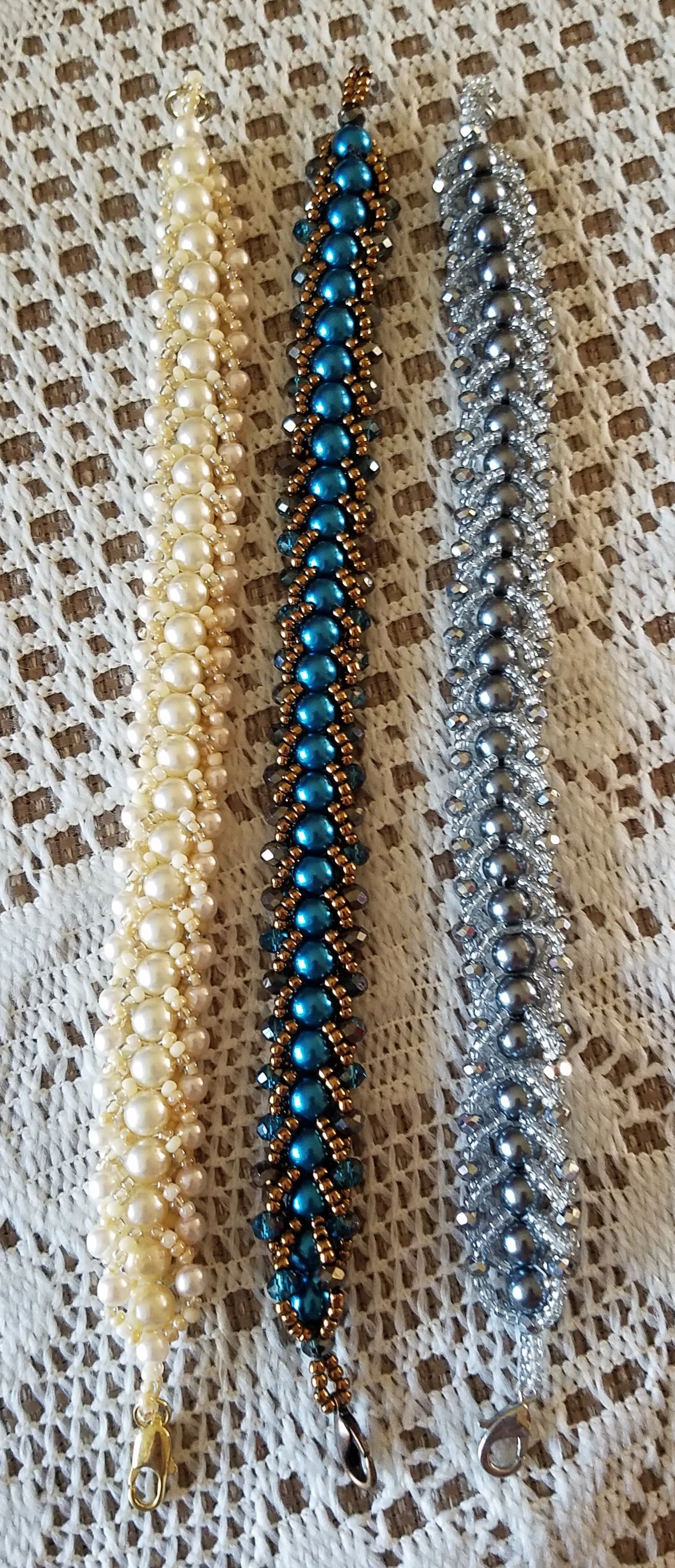 Three beaded bracelets in white, blue, and silver, laid on a woven fabric background.