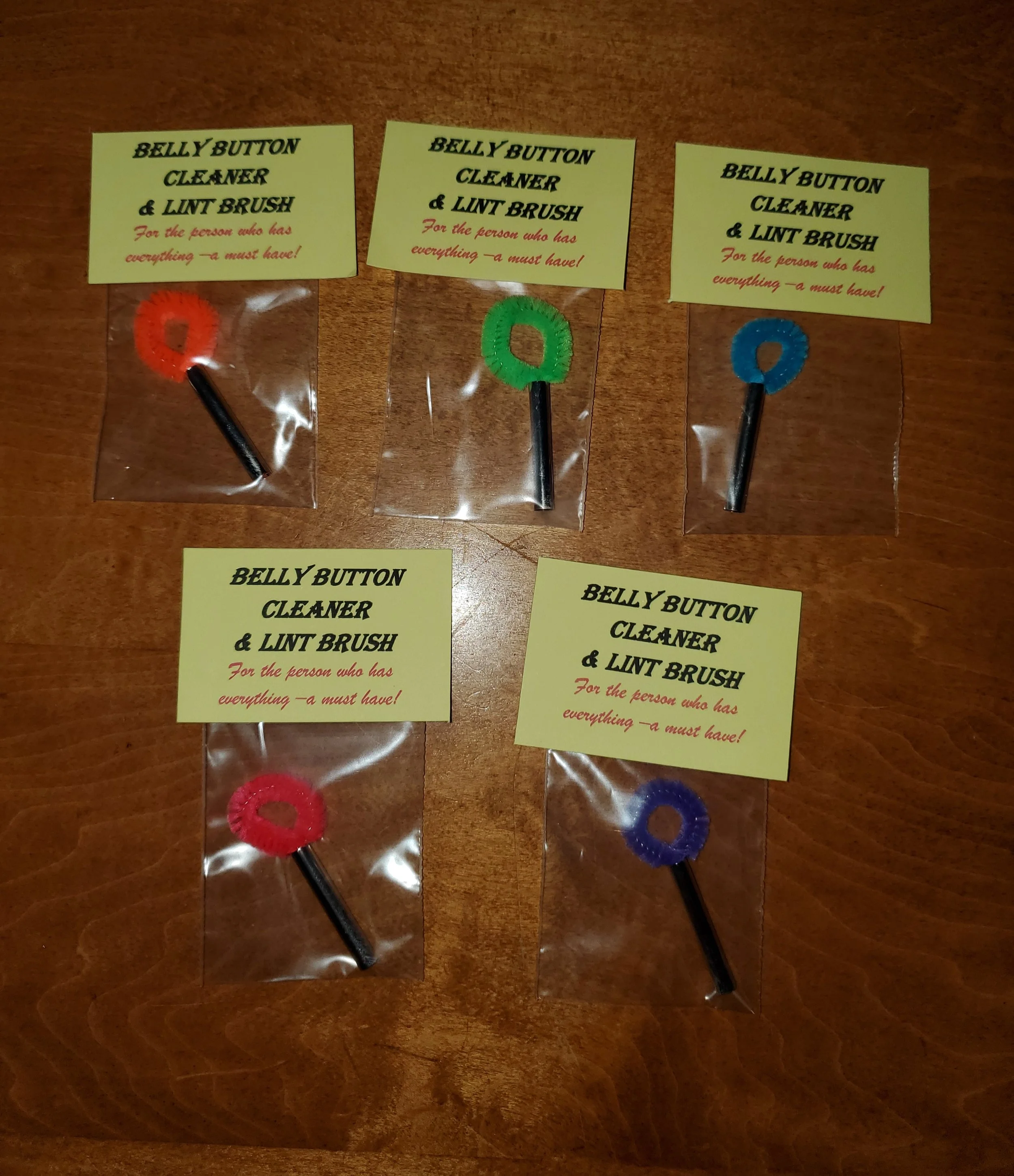 Set of five small beauty tool accessories, each with a different colored ring and a black stick, packaged in clear plastic. These accessories are labeled as 'Belly Button Cleaner & Lint Brush' with a yellow note that says, 'For the person who has eve