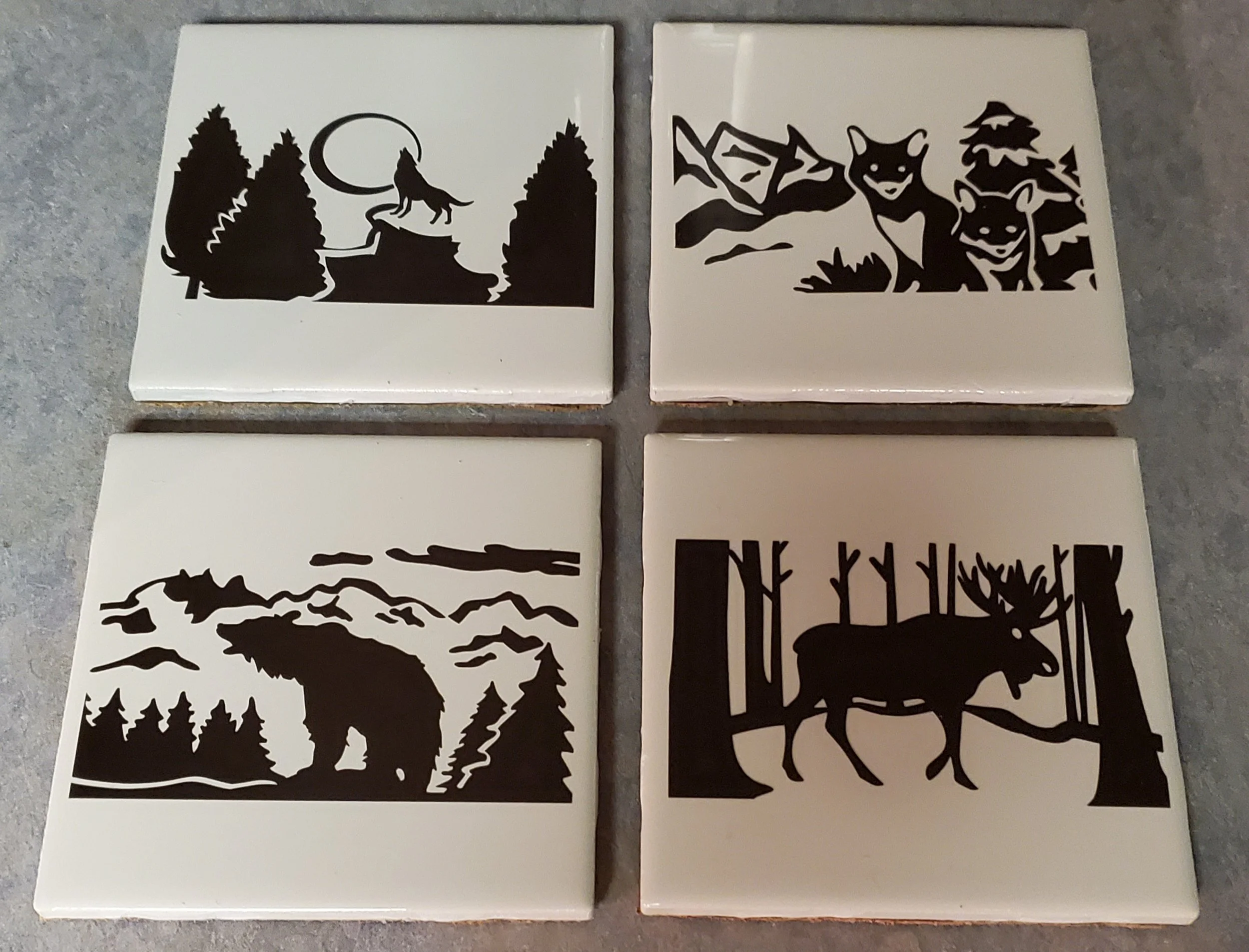 Four square tiles with black silhouette designs of woodland animals and nature scenes on a white background. Top left tile depicts a howling wolf and a full moon behind trees; top right tile shows several foxes; bottom left tile features a bear in a 