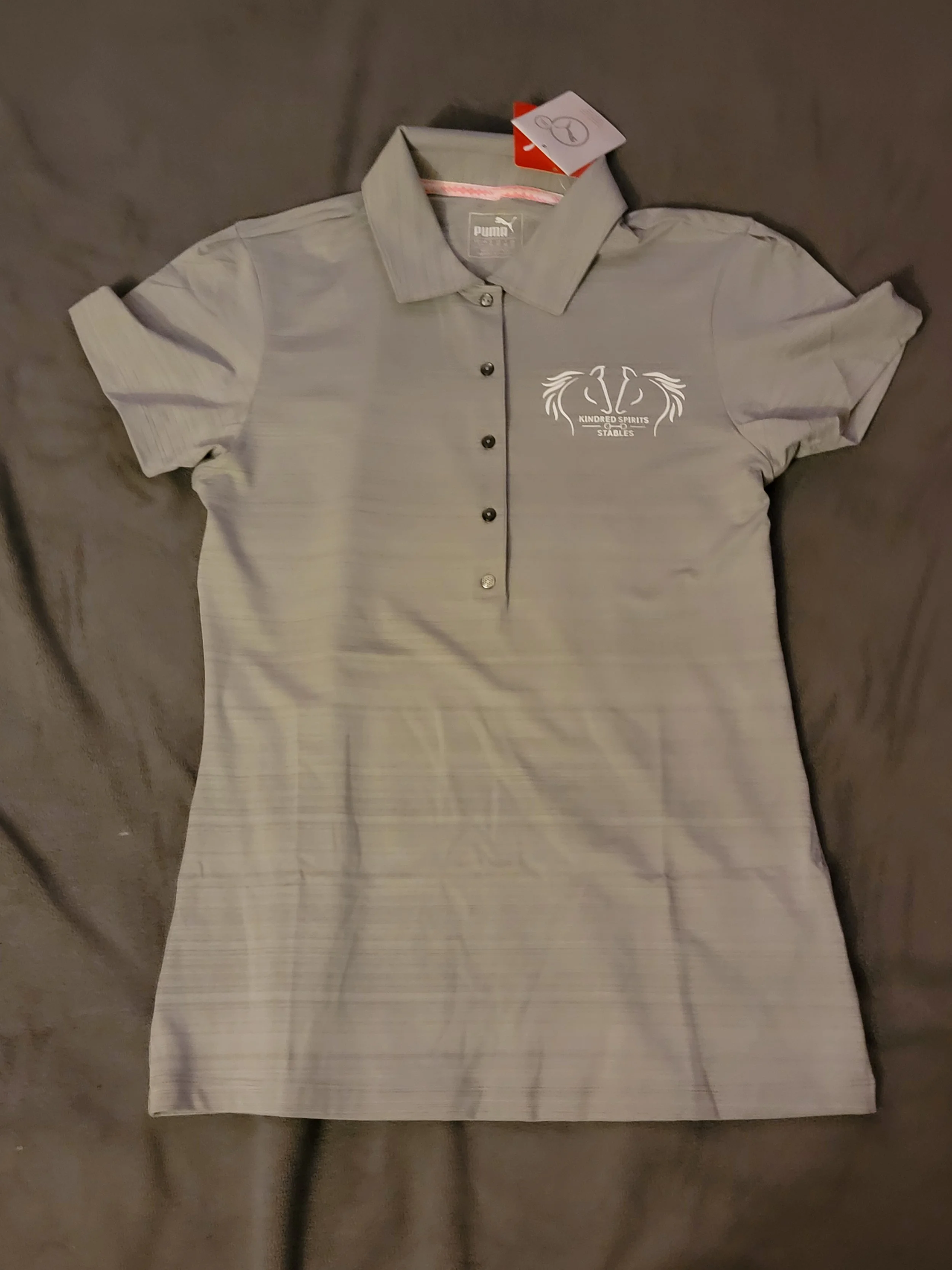 Gray polo shirt with a logo on the left chest reading 'Kindred Spirits Stables' and a stylized swan design, laid on a dark surface.