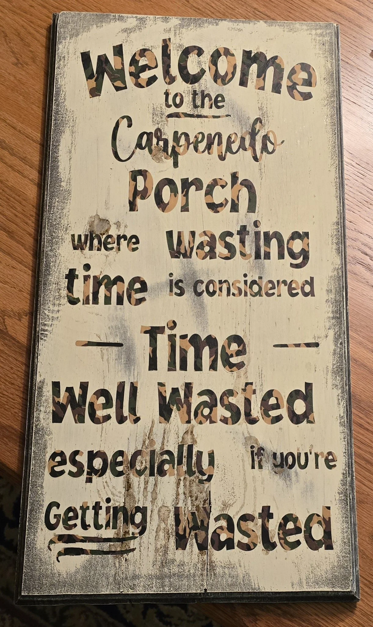 A decorative wooden sign that reads "Welcome to the Carpenter Porch where wasting time is considered time well wasted, especially if you're getting Wasted," with distressed paint and camouflage-themed lettering.
