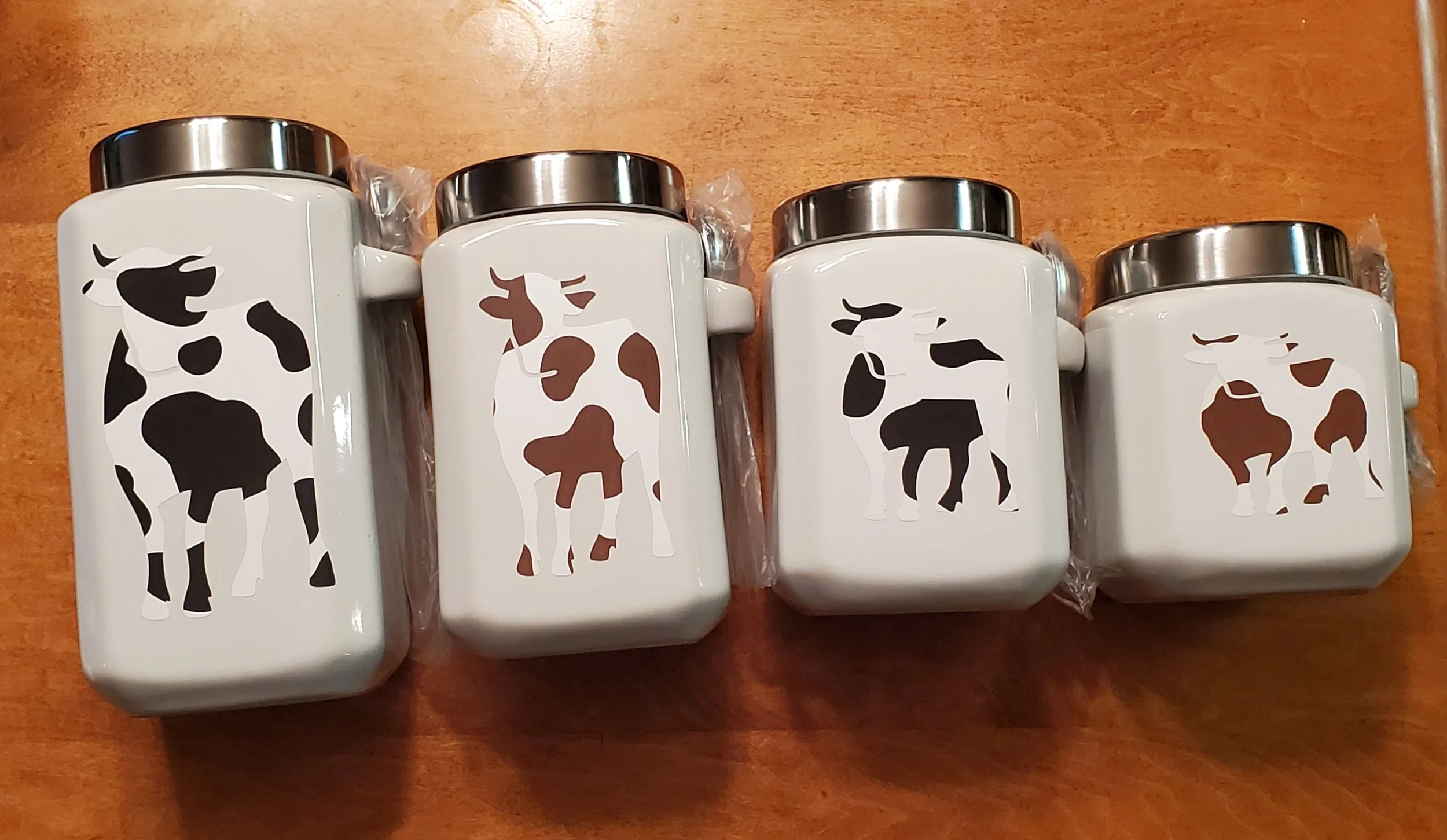 Four white ceramic canisters with silver lids featuring cow illustrations in black, brown, and white on the front, placed on a wooden surface.