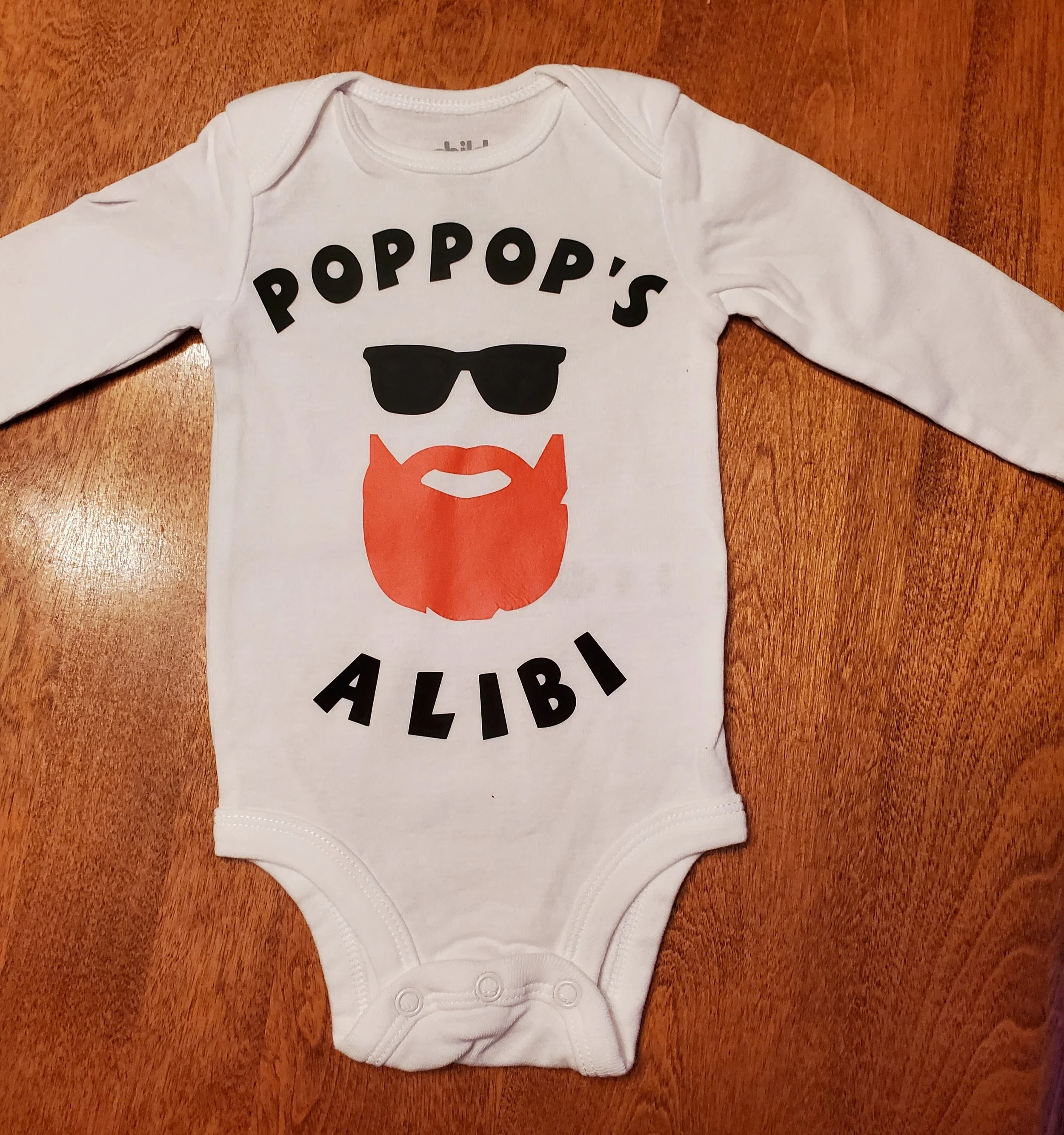 White baby onesie with black text that says 'POPPOP'S ALIBI,' featuring a graphic of sunglasses, a beard, and a mustache.