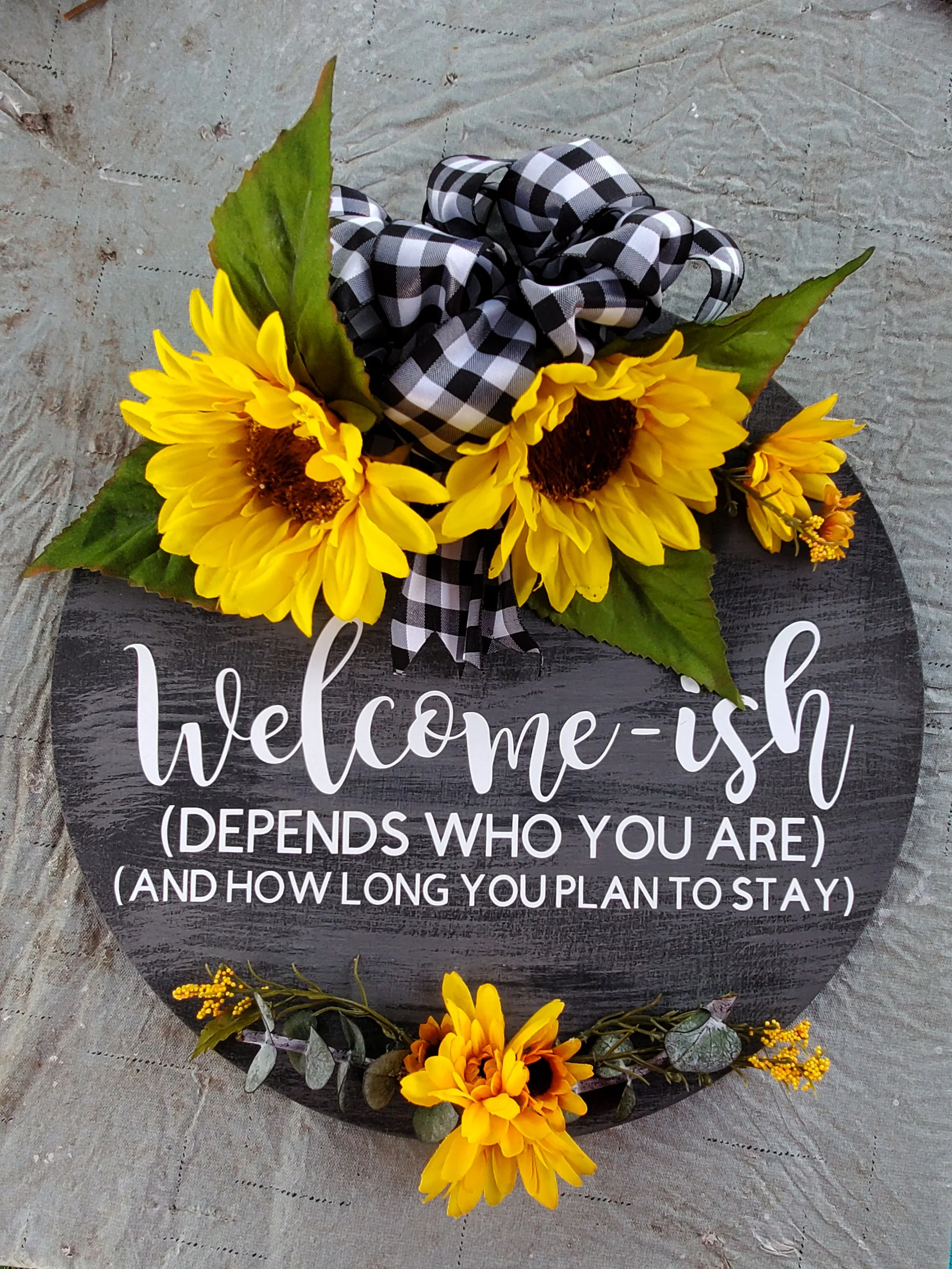 A decorative sign with sunflowers, green leaves, and a black and white checkered ribbon. The sign reads: "welcome-ish (depends who you are) and how long you plan to stay."