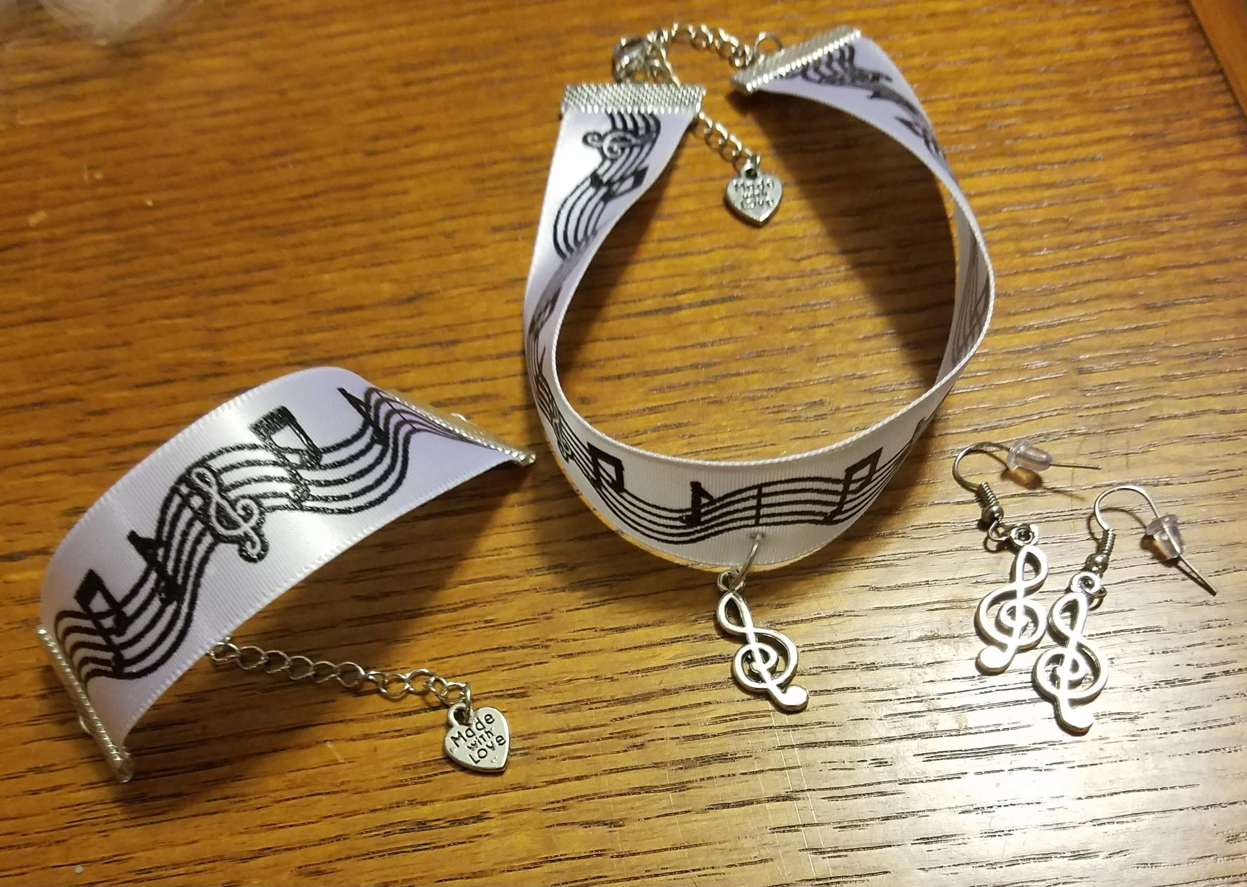 Music-themed jewelry set including a choker, earrings, and a bracelet with black musical notes and treble clefs on white ribbon and silver accents, displayed on a wooden surface.