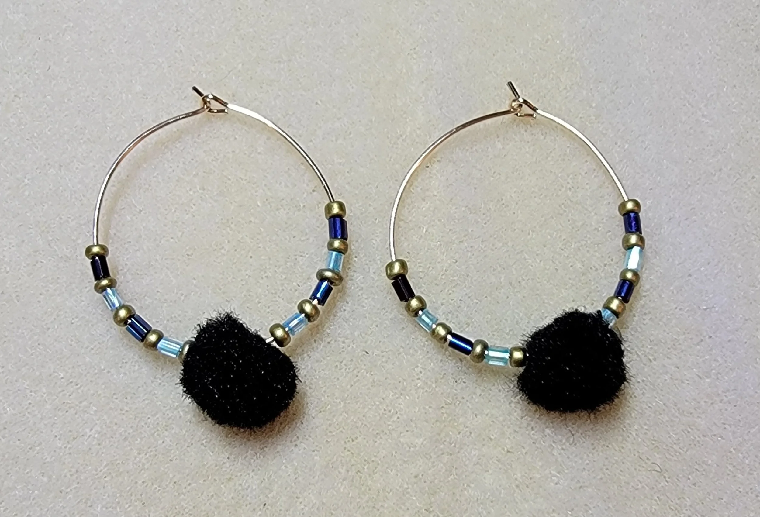 Pair of hoop earrings with blue, gold, and black beads, featuring black pom-poms at the bottom.