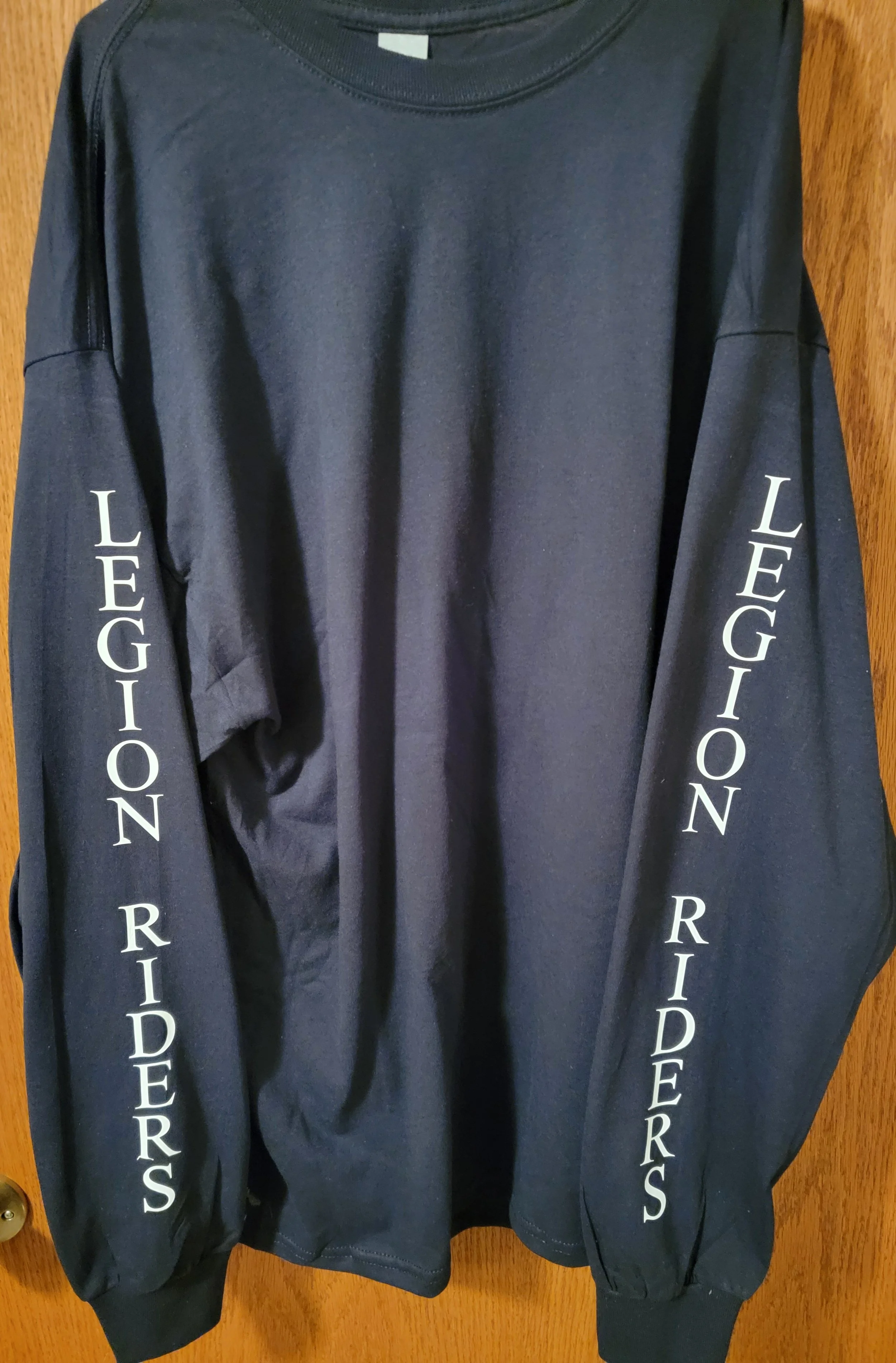 A black long-sleeve shirt hanging on a wooden door, with the words 'LEGION RIDERS' printed in white on each sleeve.