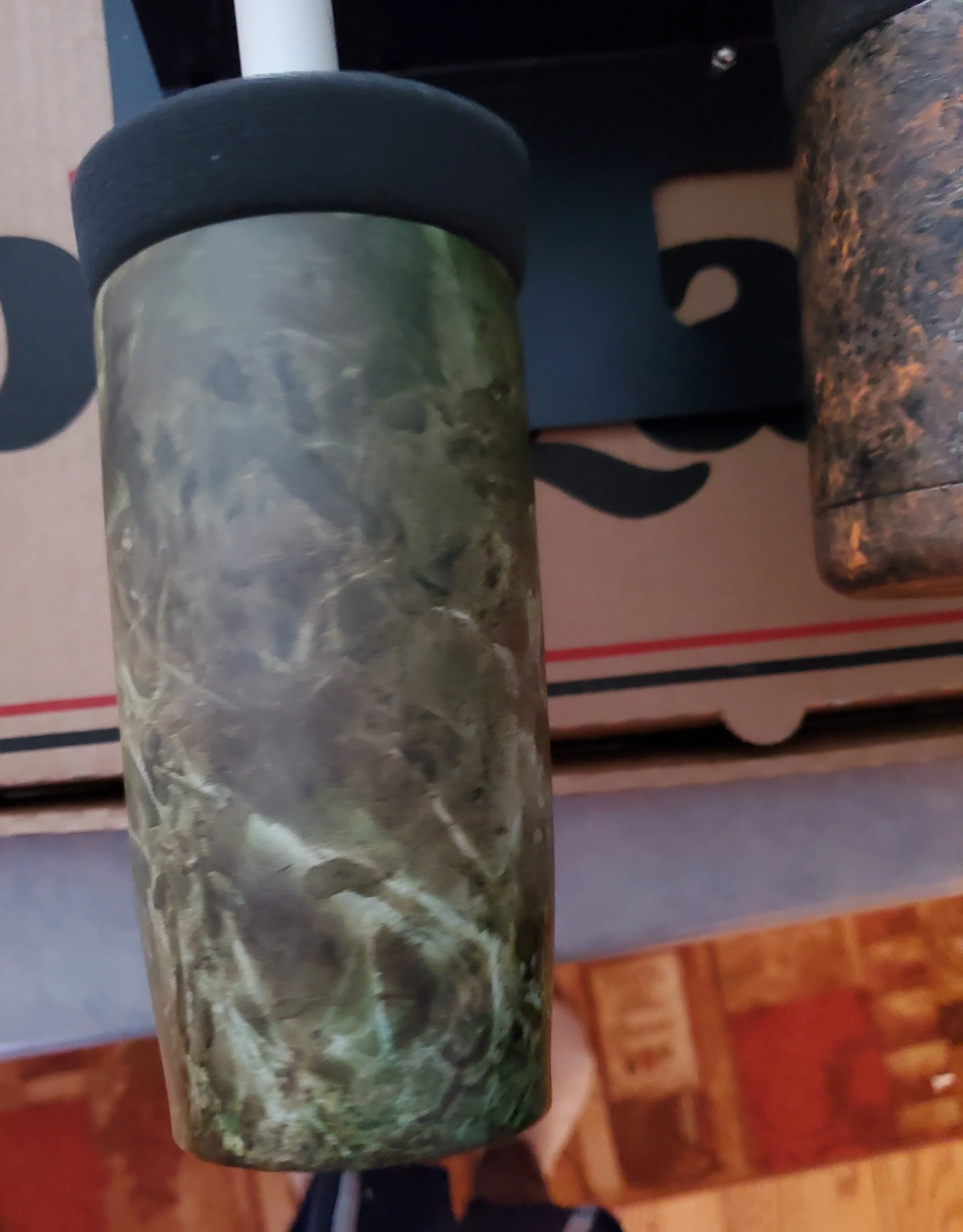 A cylindrical container with a marbled green and beige pattern, black lid, placed on a surface with cardboard boxes and other objects nearby.