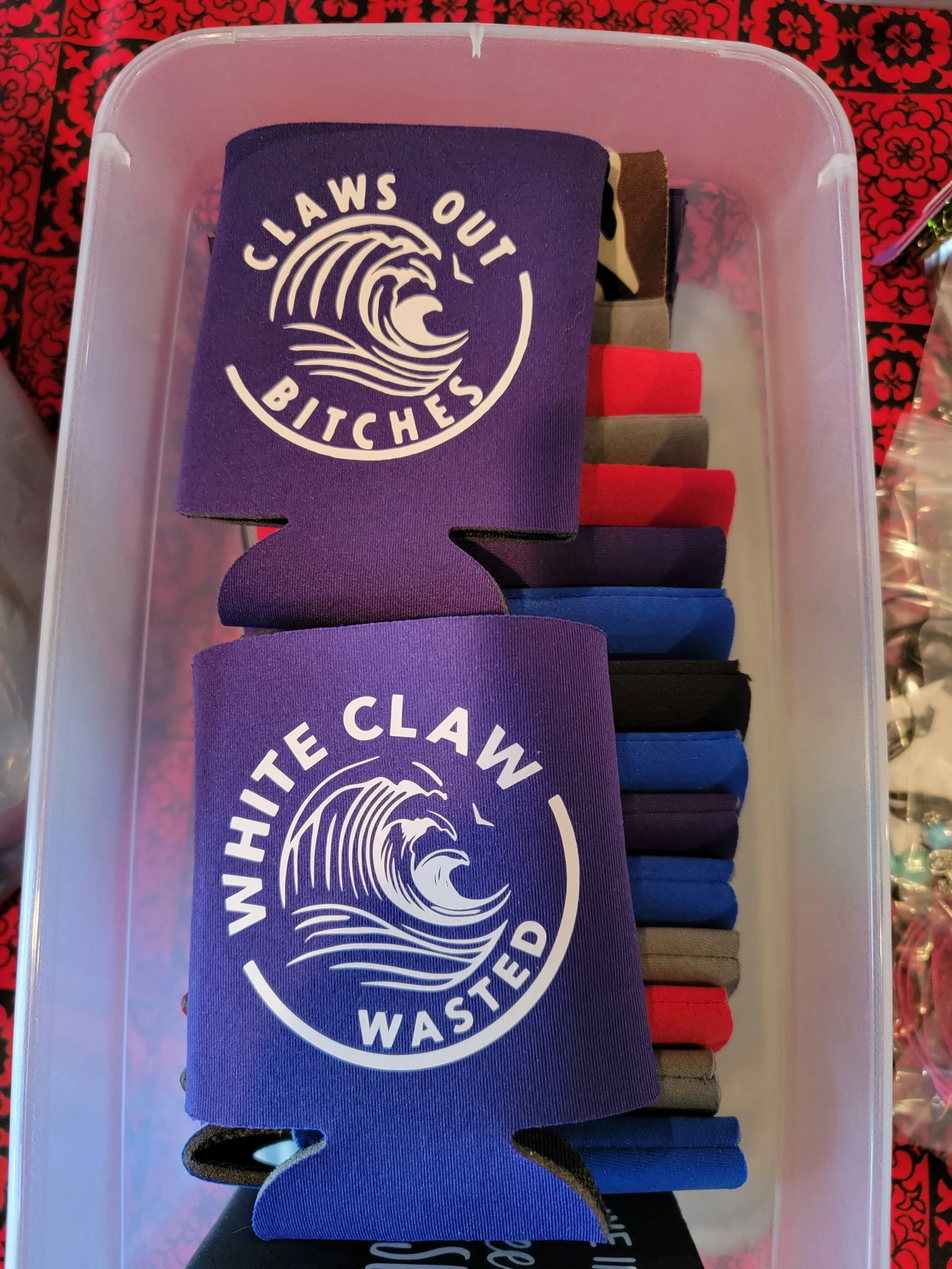 A plastic storage bin filled with assorted colorful fabric wristbands, some with the phrase "Claws Out Bitches" and others with the phrase "White Claw Wasted," featuring a wave illustration.