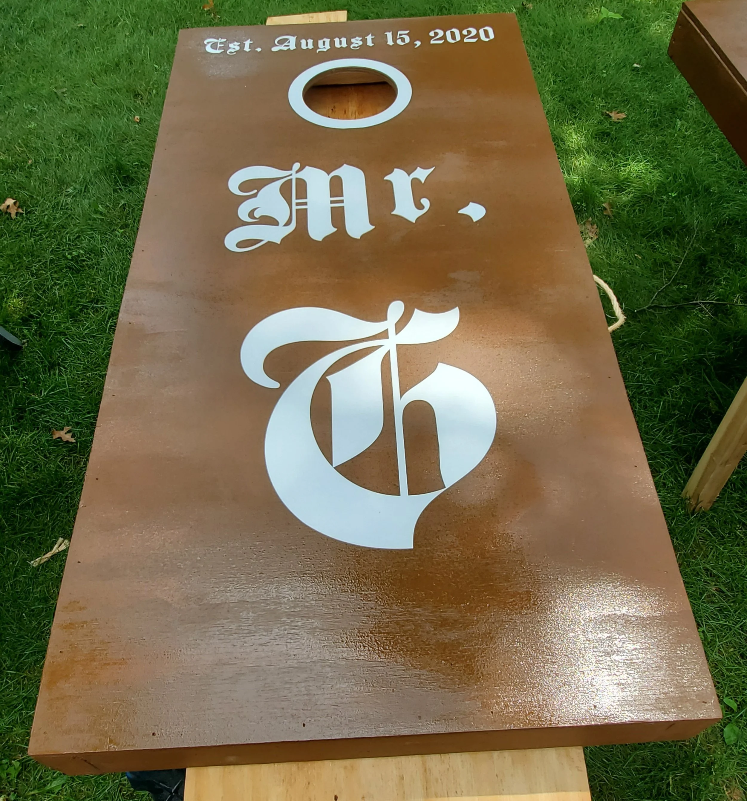 A cornhole board with the text 'Est. August 15, 2020' at the top, and a large white letter 'M' with a decorative flourish in the center, placed outdoors on a grassy area.