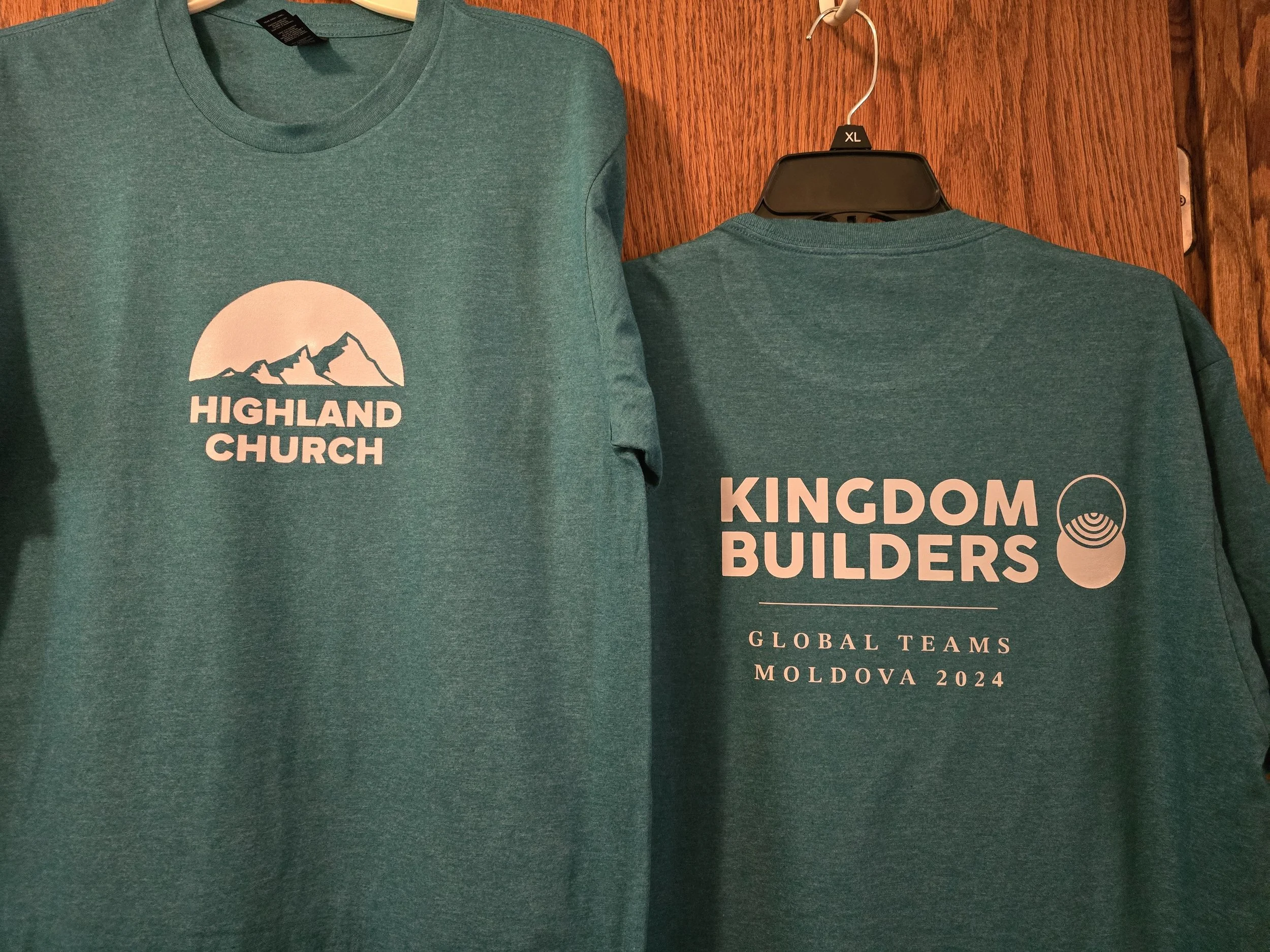 Two teal-colored T-shirts hanging on a wooden wall; the front shirt has a mountain logo with the text 'Highland Church,' and the back shirt has the text 'Kingdom Builders,' along with a logo and additional text mentioning 'Global Teams Moldova 2024.'