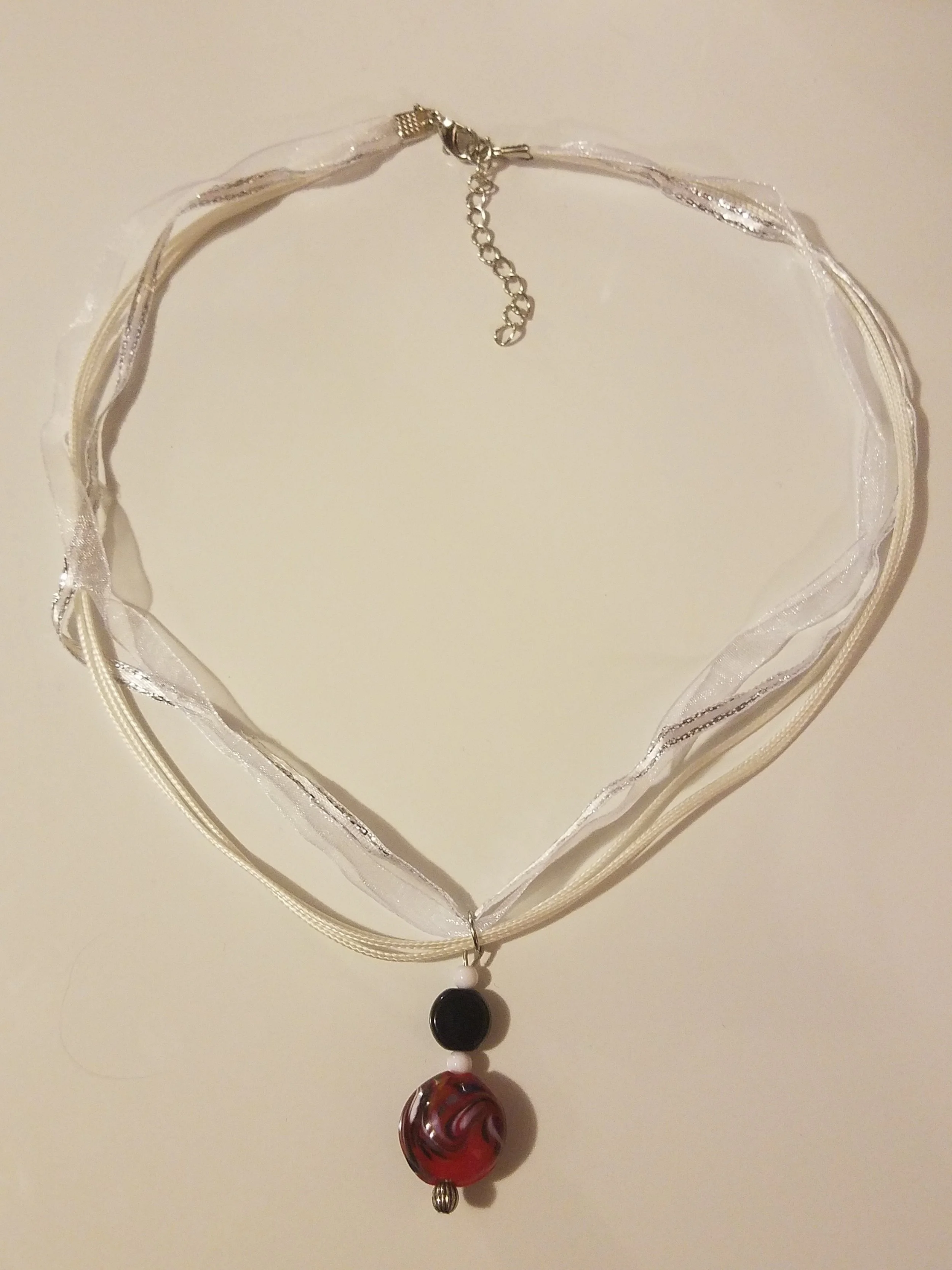 A layered necklace with a black bead, a white bead, and a red and black swirled bead, on a white fabric ribbon with silver accents.