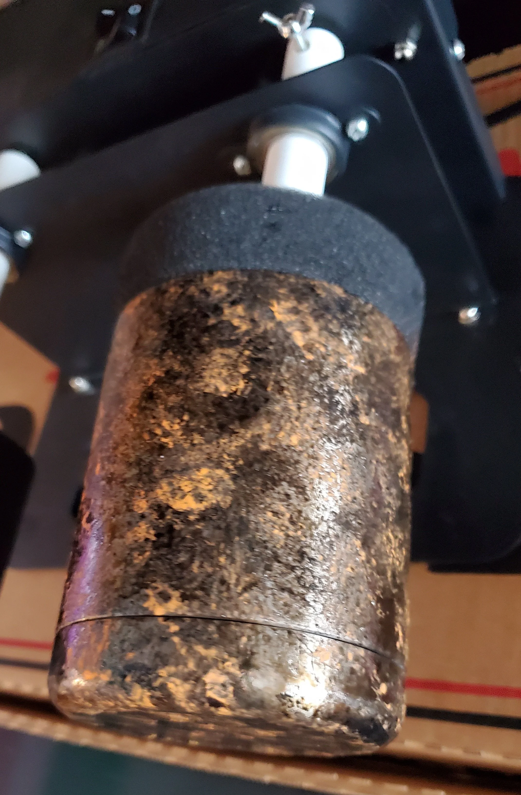 Close-up of a rusted, muddy cylindrical object attached to a black mechanical structure with white bolts and a black foam cover on top.
