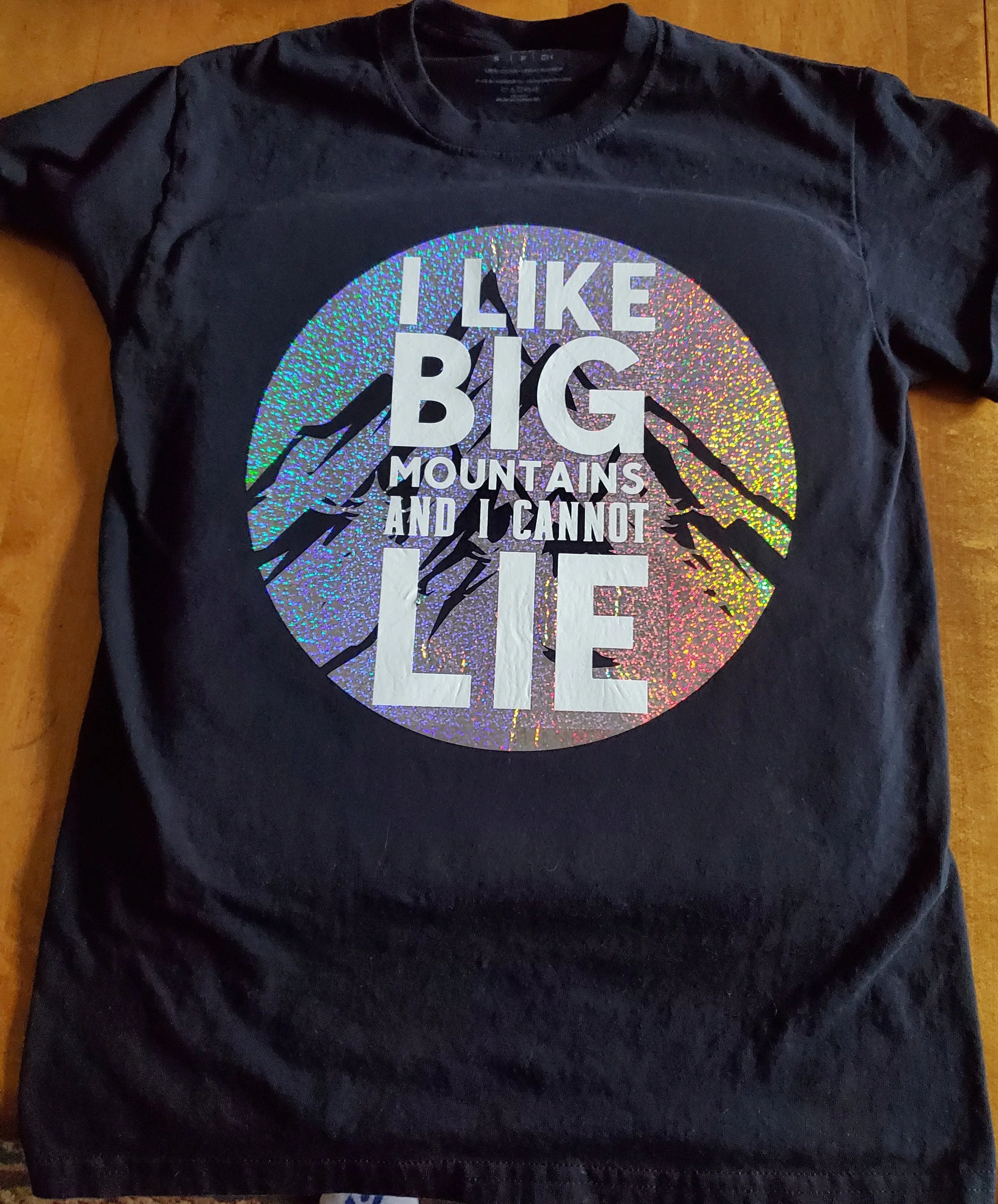 Black T-shirt with holographic print of mountain peaks and the phrase 'I like big mountains and I cannot lie' in large white text.