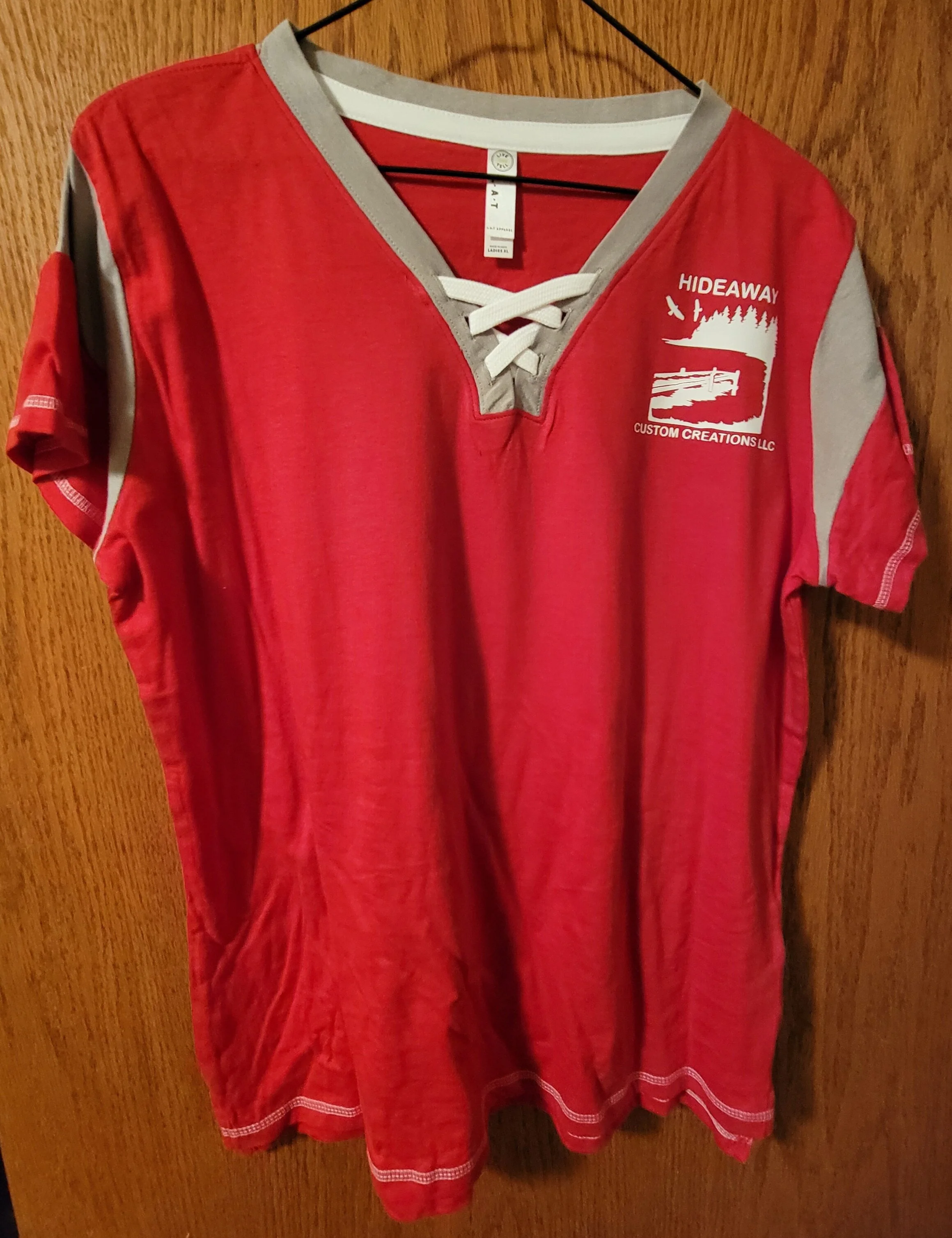 Red short-sleeve hockey jersey with gray accents, lace-up V-neck, and a logo on the left chest reading 'HIDEAWAY CUSTOM CREATIONS LLC' with a graphic of a forest and lake scene.