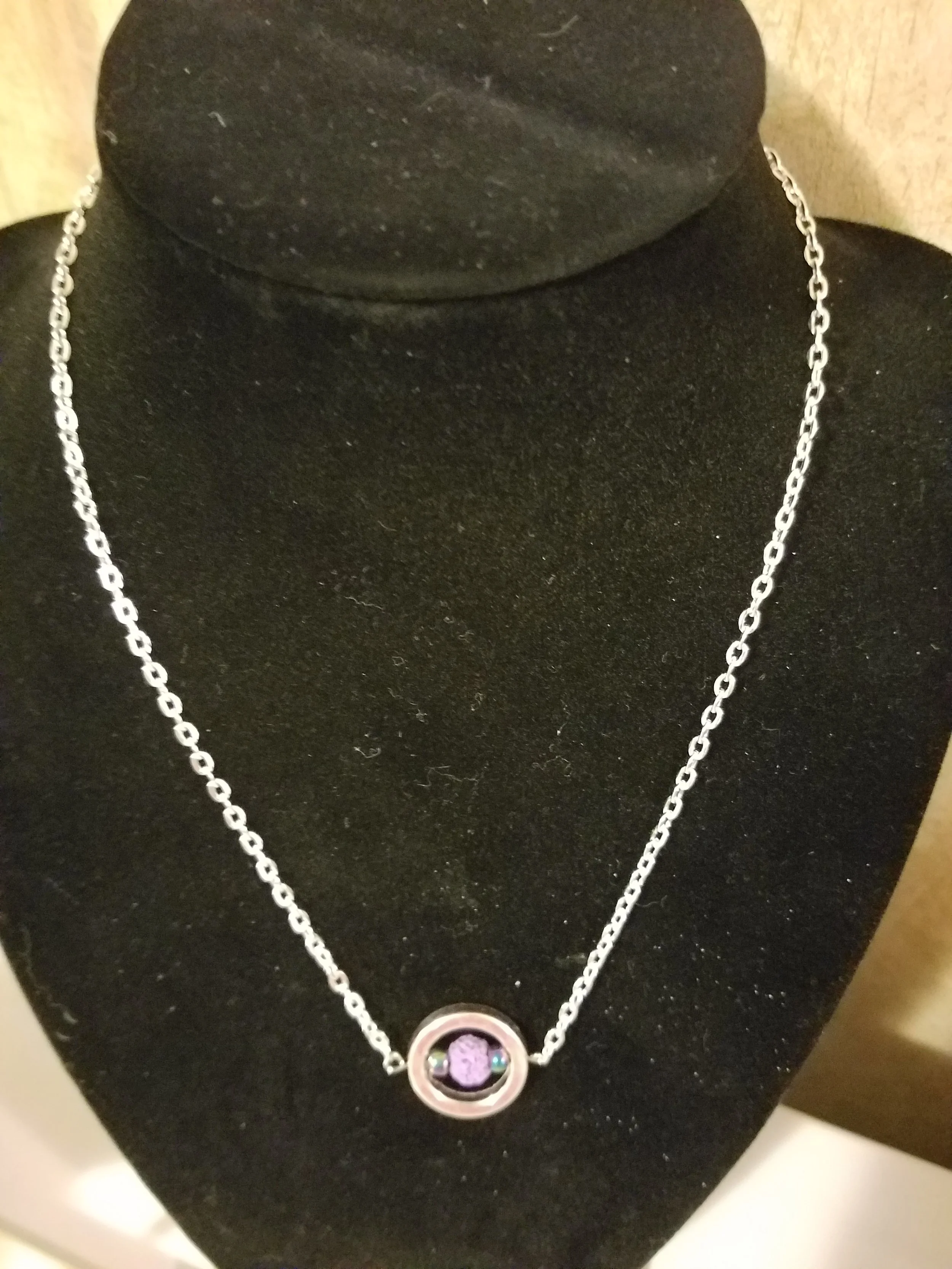 Silver necklace with a round pendant containing a purple gemstone, displayed on a black jewelry bust.