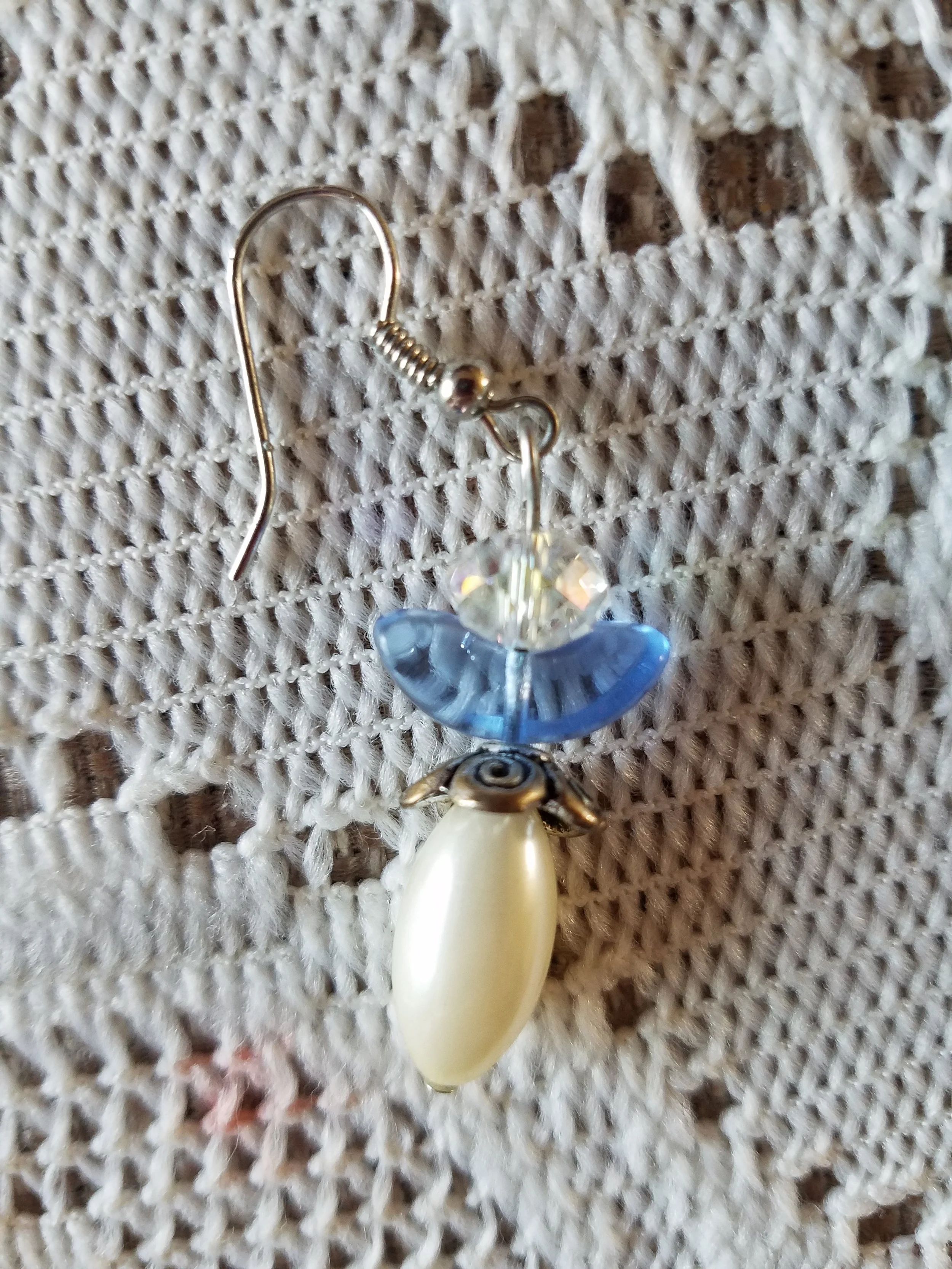 Earrings with a shell, a blue curved bead, and a clear faceted bead, hanging on a textured beige fabric.