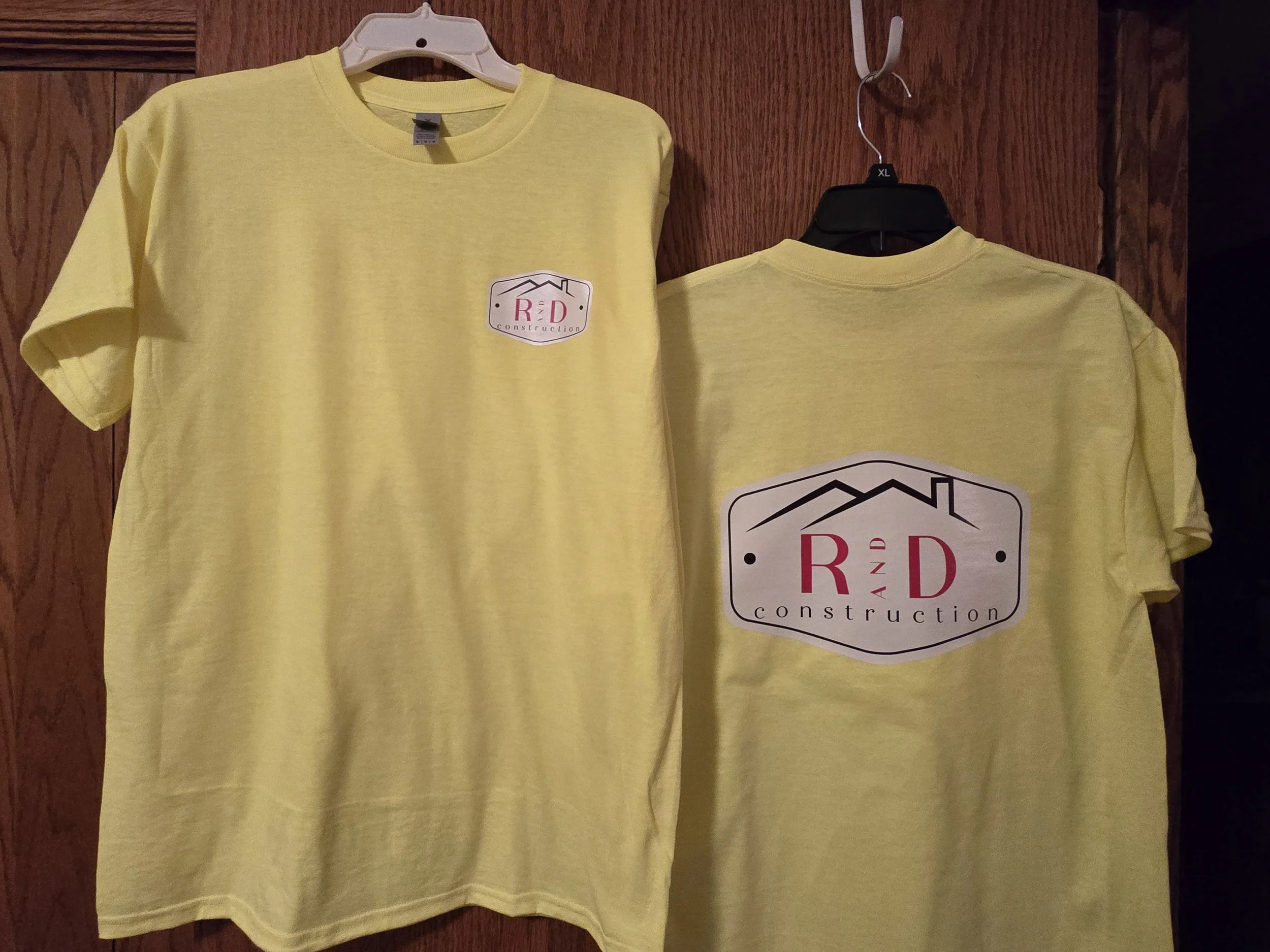 Two yellow T-shirts with a logo that says 'R&D Construction' hanging on wooden hooks.