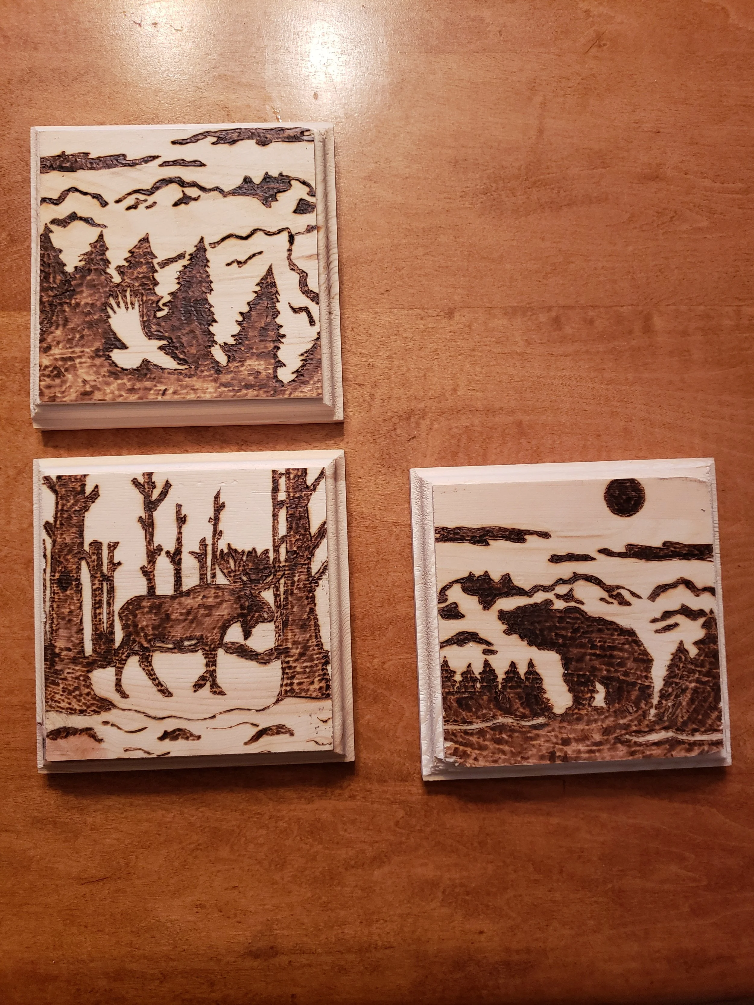 Three wooden coasters with laser-engraved nature scenes, including trees, mountains, and wildlife, on a wooden surface.