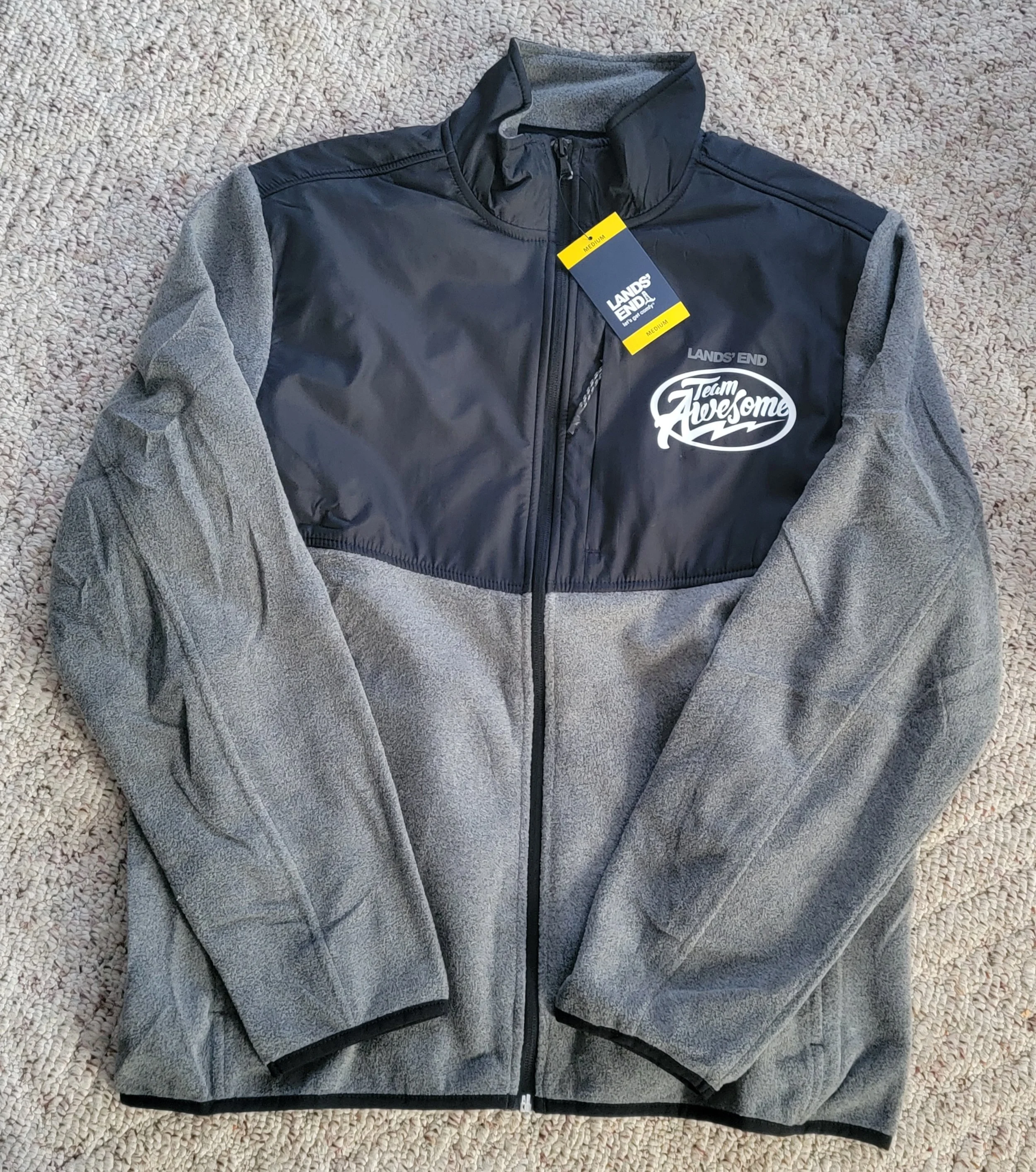 Gray and black zip-up fleece jacket with tags and a logo reading 'Team Awesome' on the front, laid on a beige carpet.