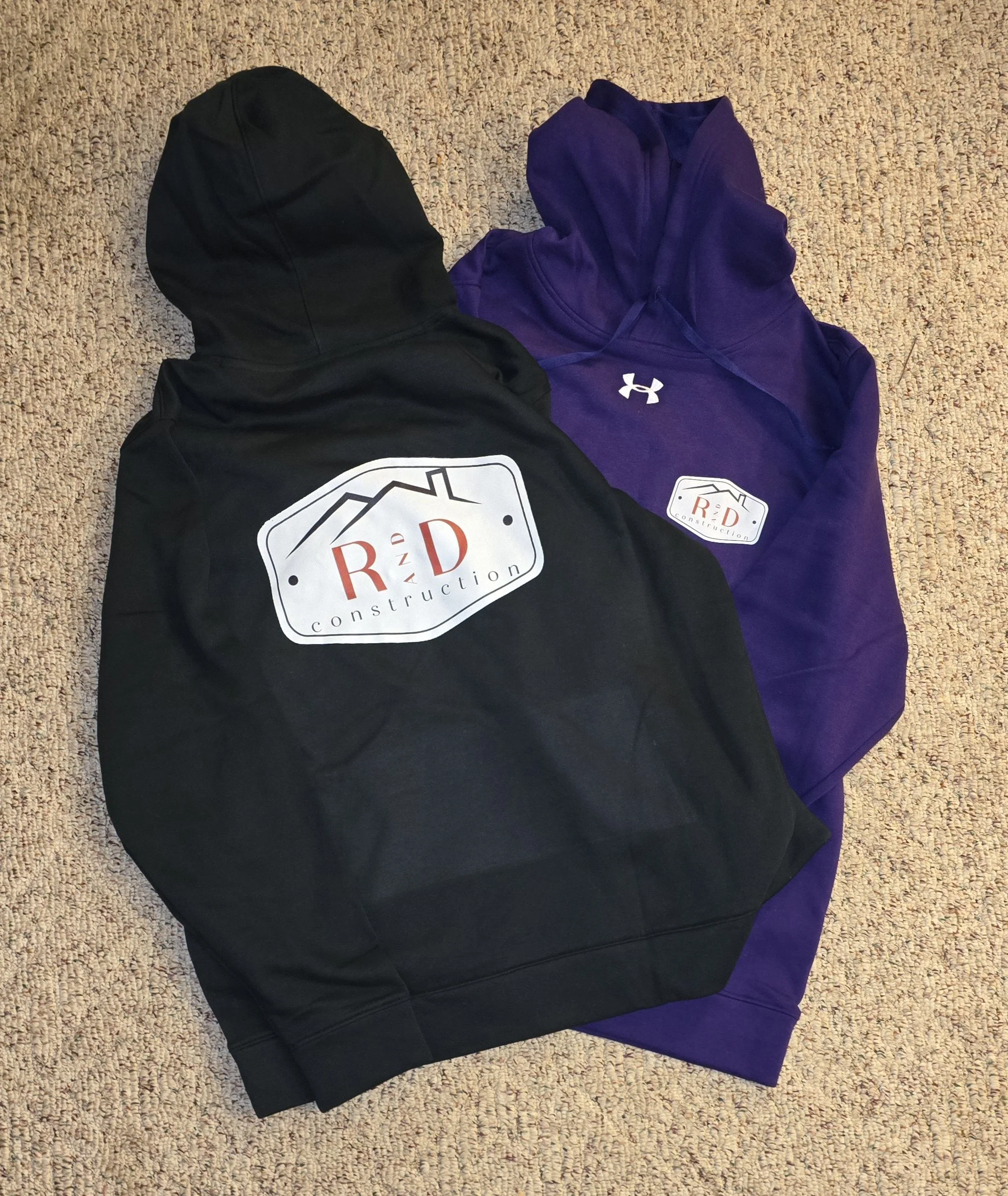 Two hoodies, one black and one purple, with logos on the front, laid out on a beige carpet.
