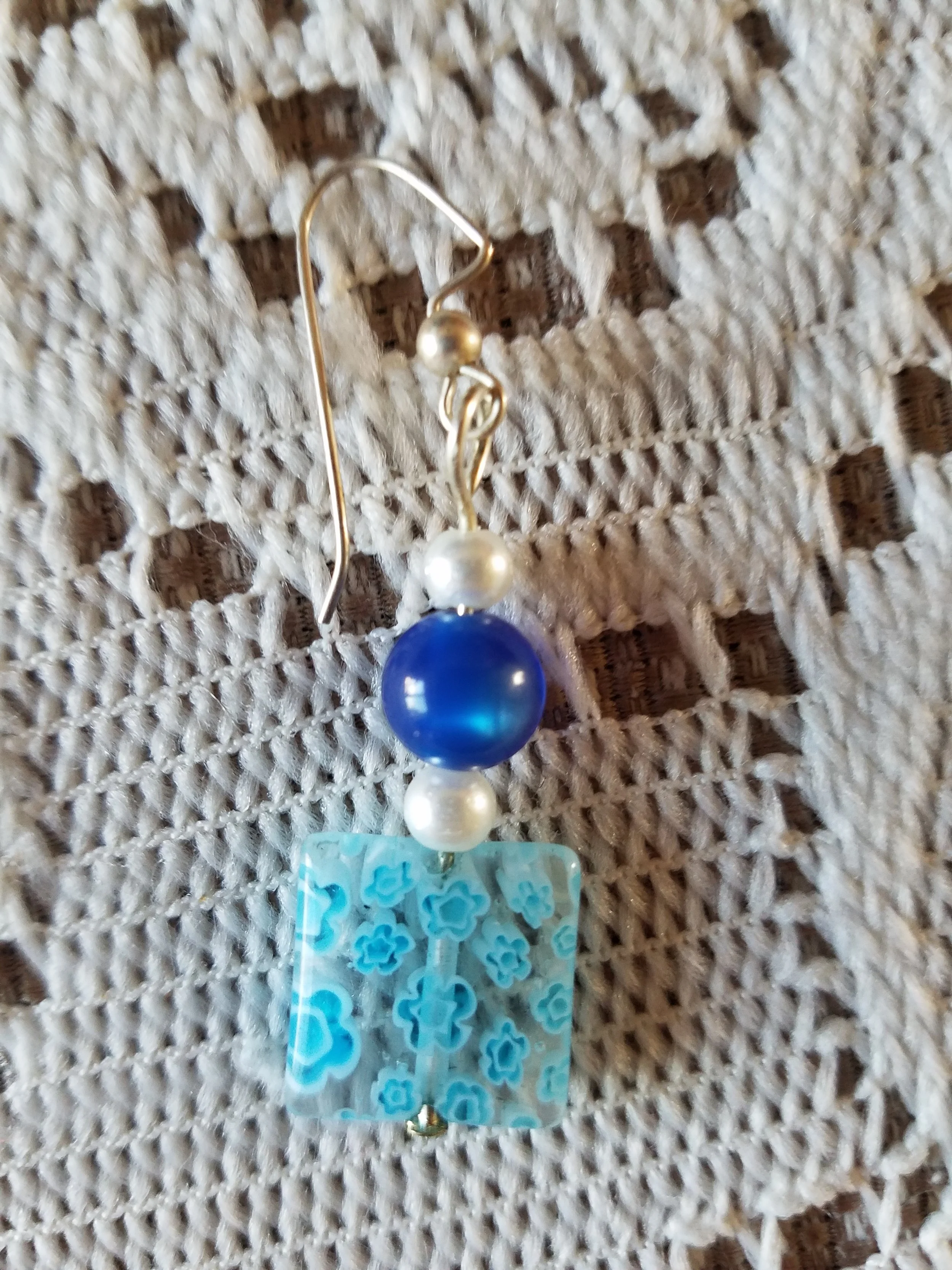 A handmade earring with a silver hook, white and blue beads, and a rectangular blue flower-patterned pendant, resting on a textured beige fabric.