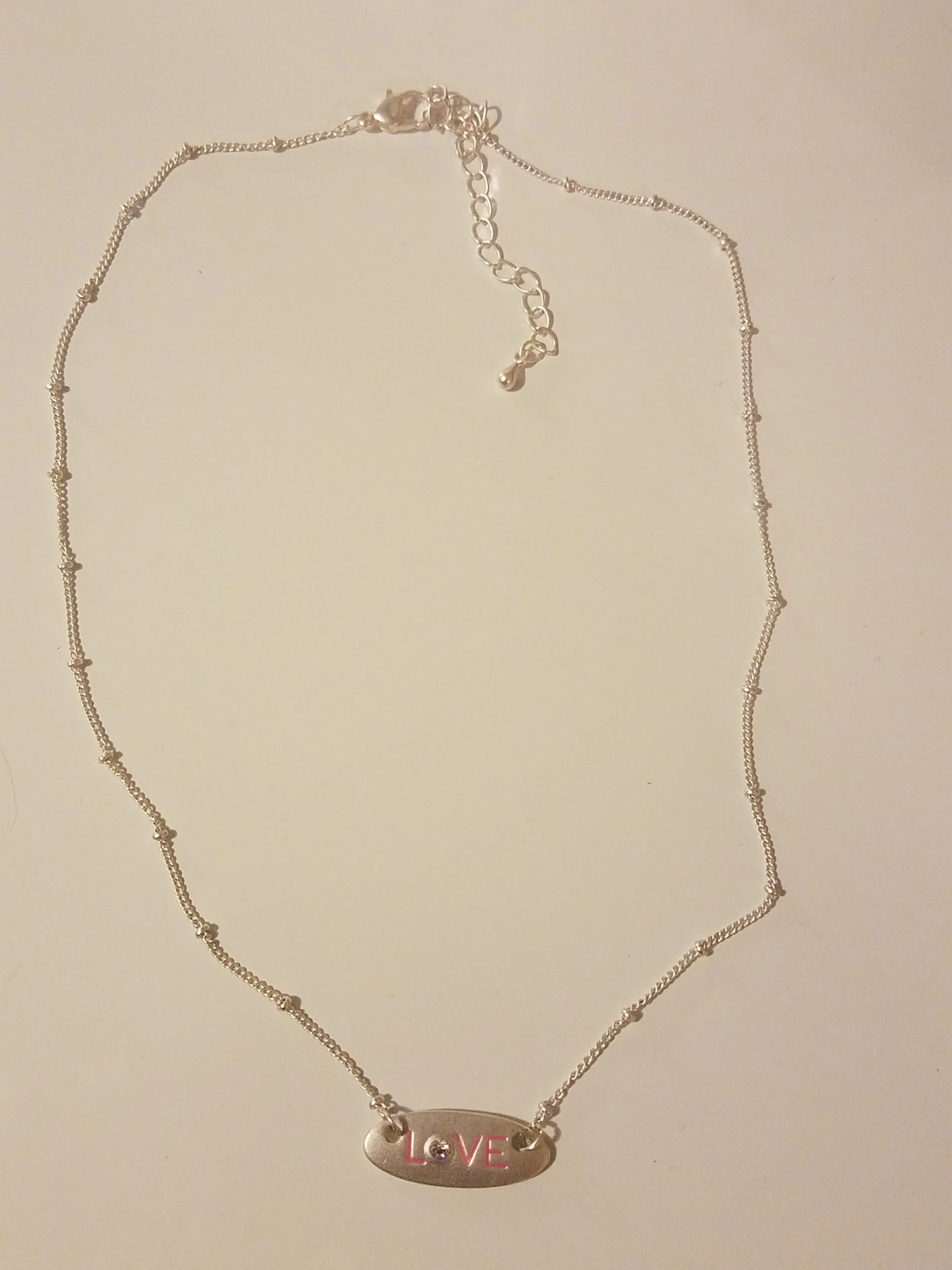 Silver necklace with a pendant spelling 'LOVE' in red with a small gemstone, laid flat on a plain surface.