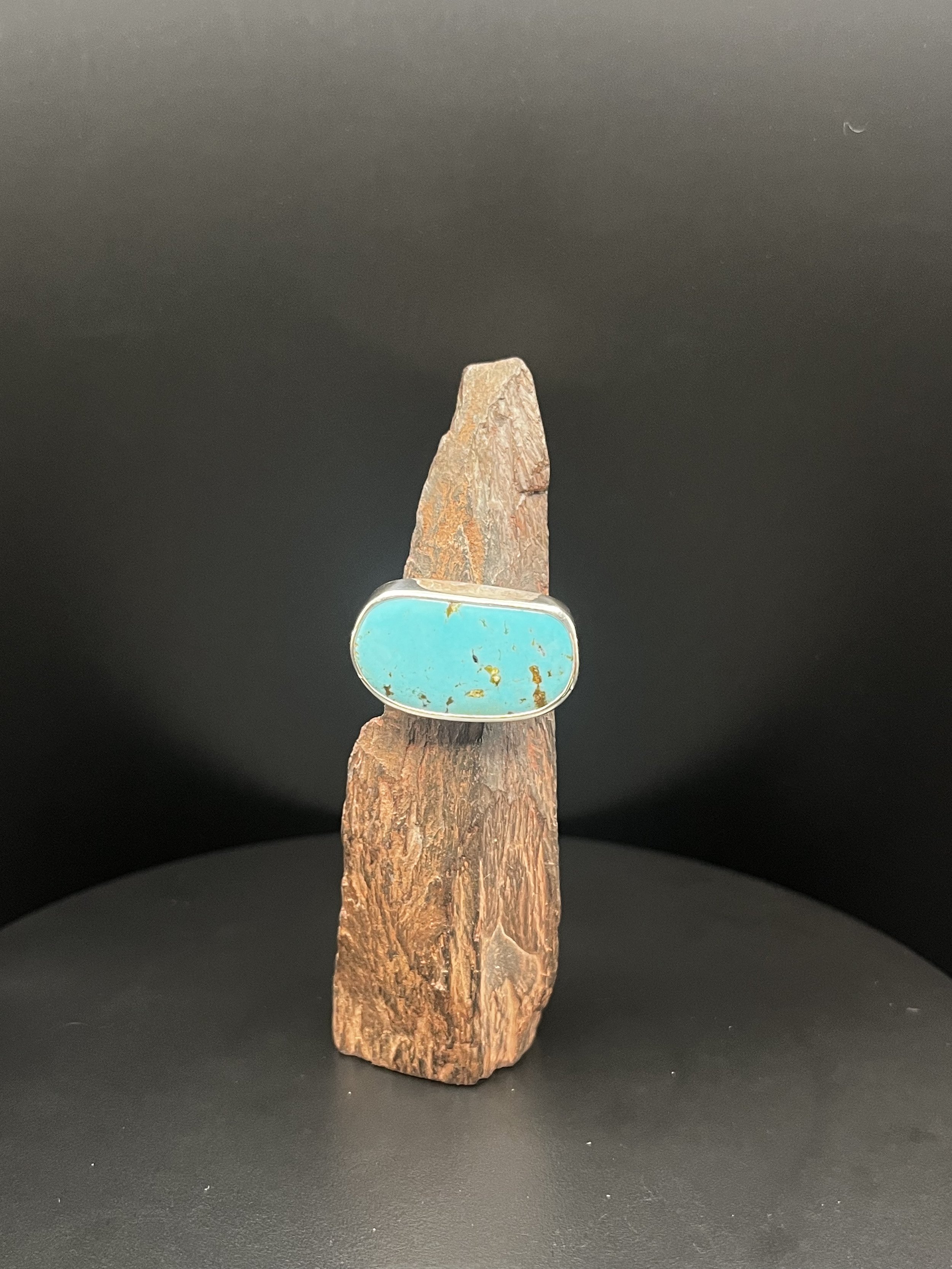 A large ring with a Kingman turquoise gemstone inside a sterling silver band, wrapped around a jagged rock rested on a black display stand, surrounded by black walls.