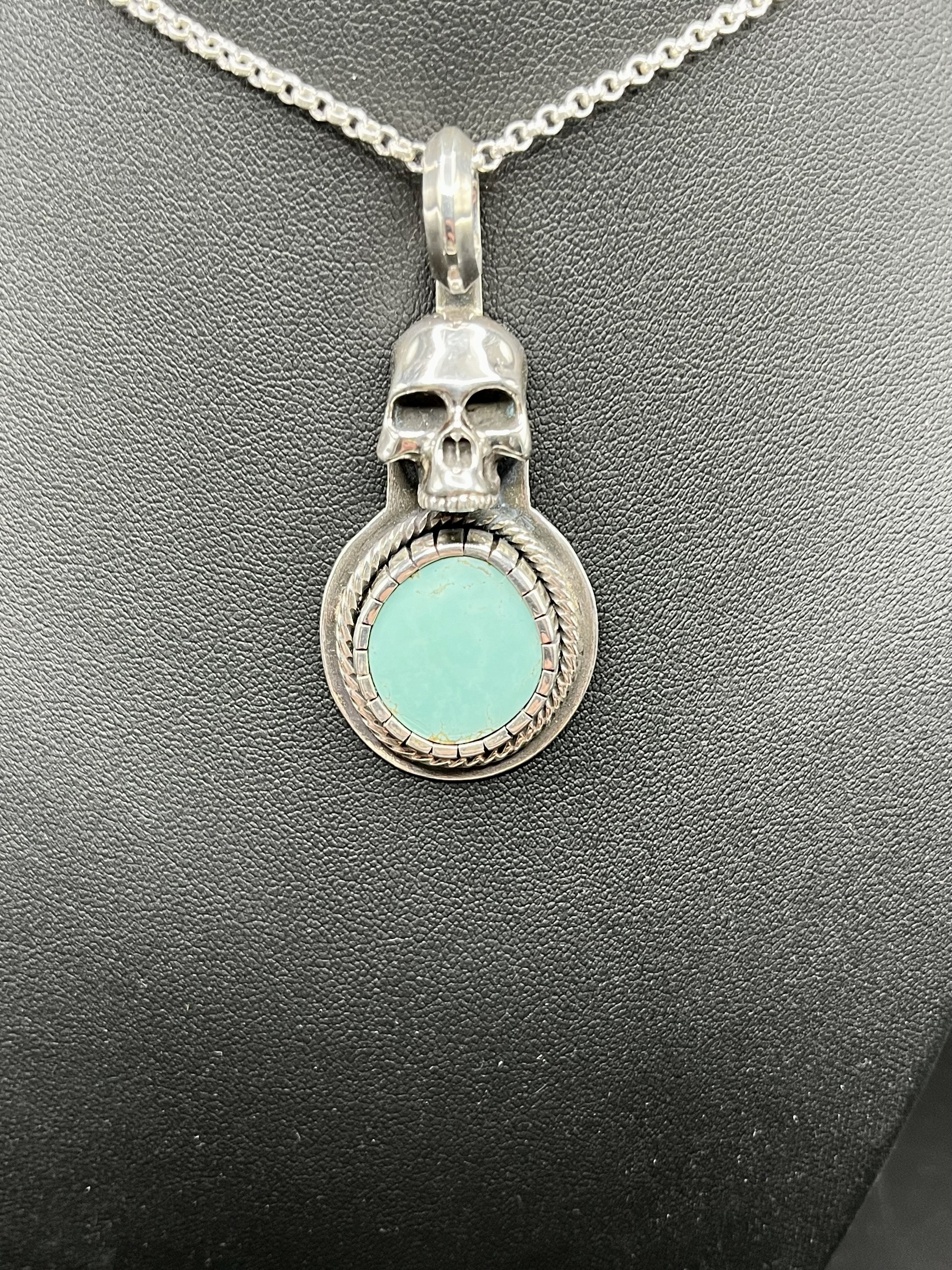 A silver necklace with a skull pendant and bright turquoise gemstone displayed on a black mannequin.