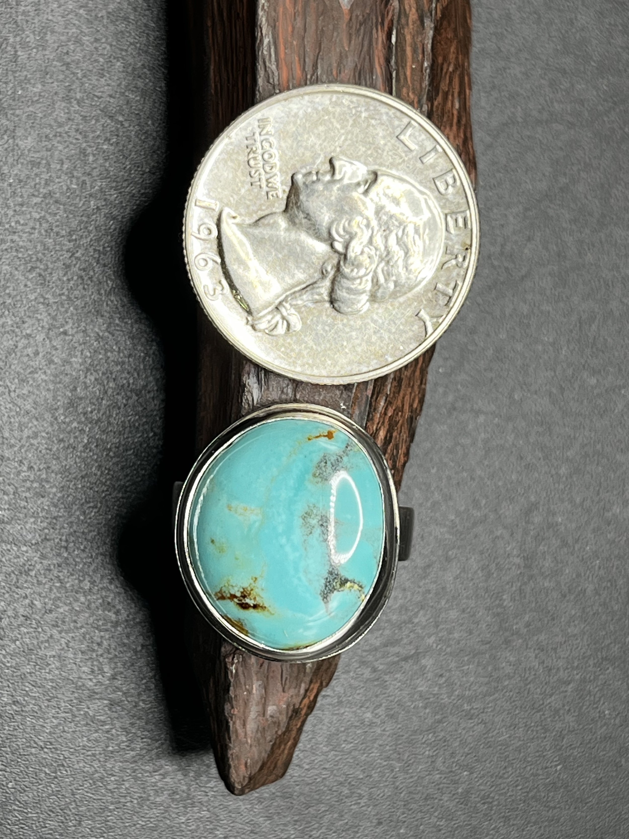 A ring with a large turquoise gemstone displayed on a jagged rock. There is a heads-up quarter rested on the rock to illustrate the size of the turquoise gemstone.