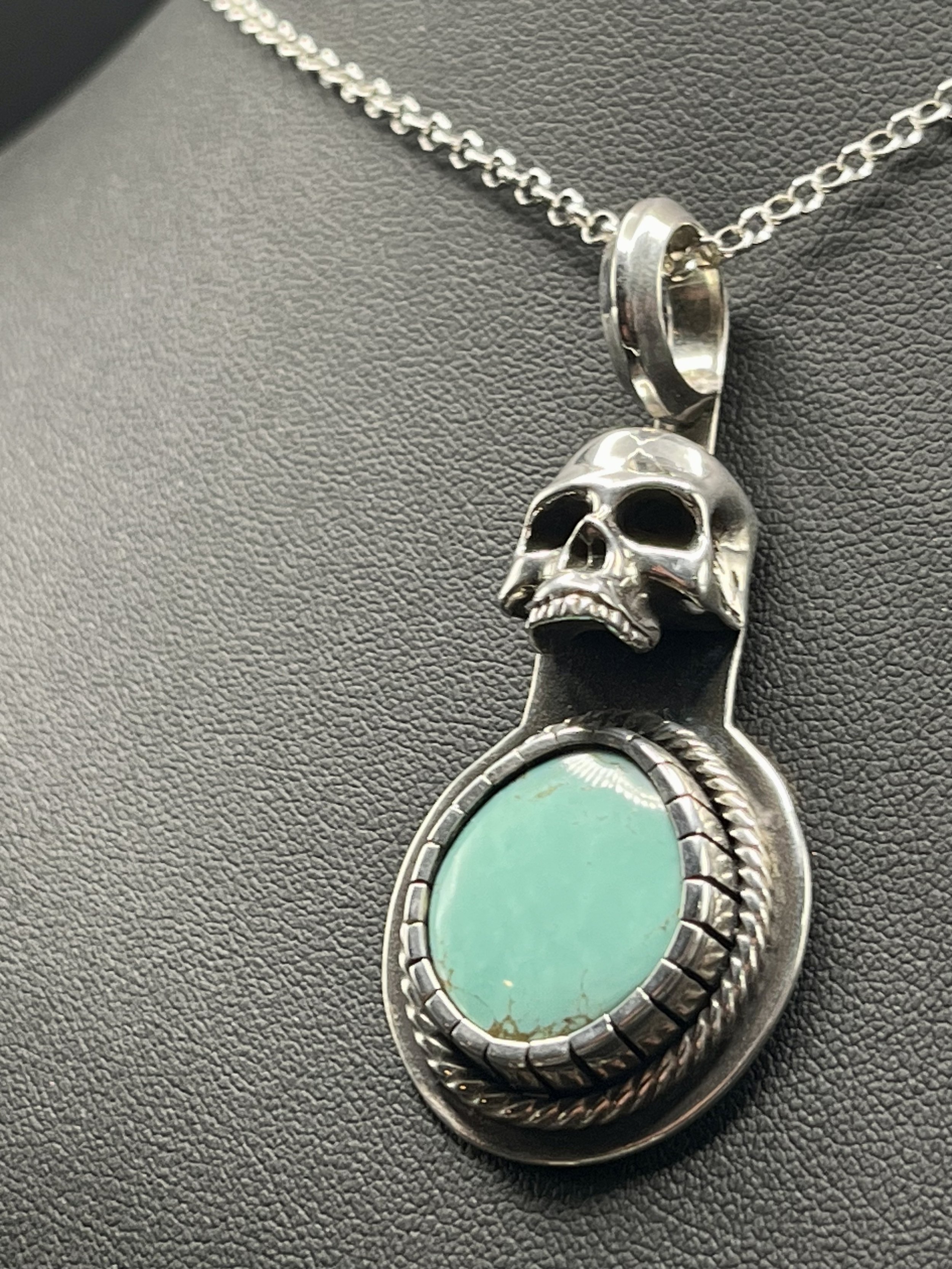 A silver necklace with a skull pendant and bright turquoise gemstone displayed on a black mannequin, angled slightly away from the camera to showcase the sterling silver chain.