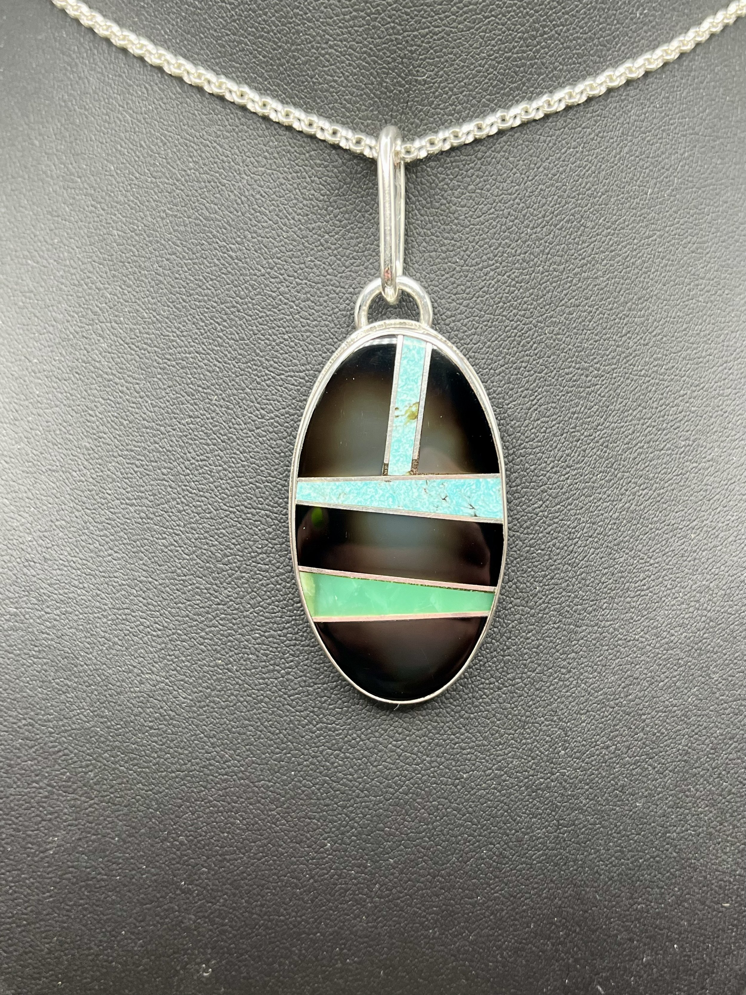 A pendant necklace from KC Silversmith with a black, blue, and green pendant displayed on a small black mannequin.
